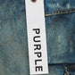 Close-up of a white rectangular tag with the words "PURPLE BRAND" in black text, attached to PURPLE BRAND P018-TPDI TRIPLE POCKET CARGO DK INDIGO premium stretch denim fabric with an orange beaded chain.