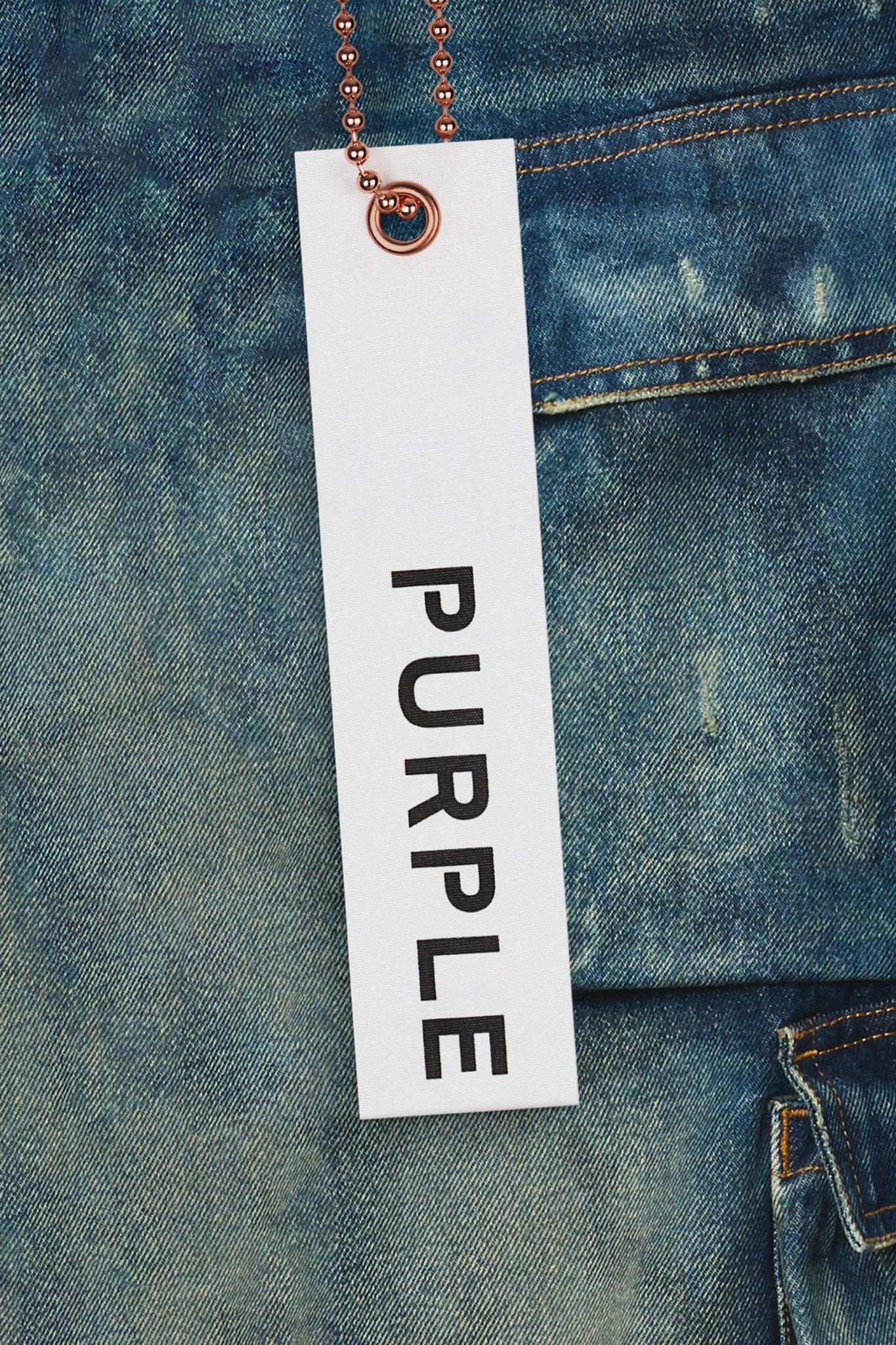 Close-up of a white rectangular tag with the words "PURPLE BRAND" in black text, attached to PURPLE BRAND P018-TPDI TRIPLE POCKET CARGO DK INDIGO premium stretch denim fabric with an orange beaded chain.