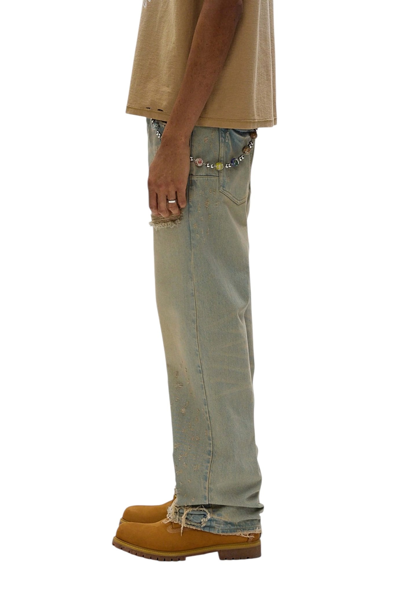 Side view of a person wearing a beige shirt, PURPLE BRAND's P018-VEDL Verditer Destroyed LT Indigo jeans with a chain belt, and brown work boots, standing against a white background.