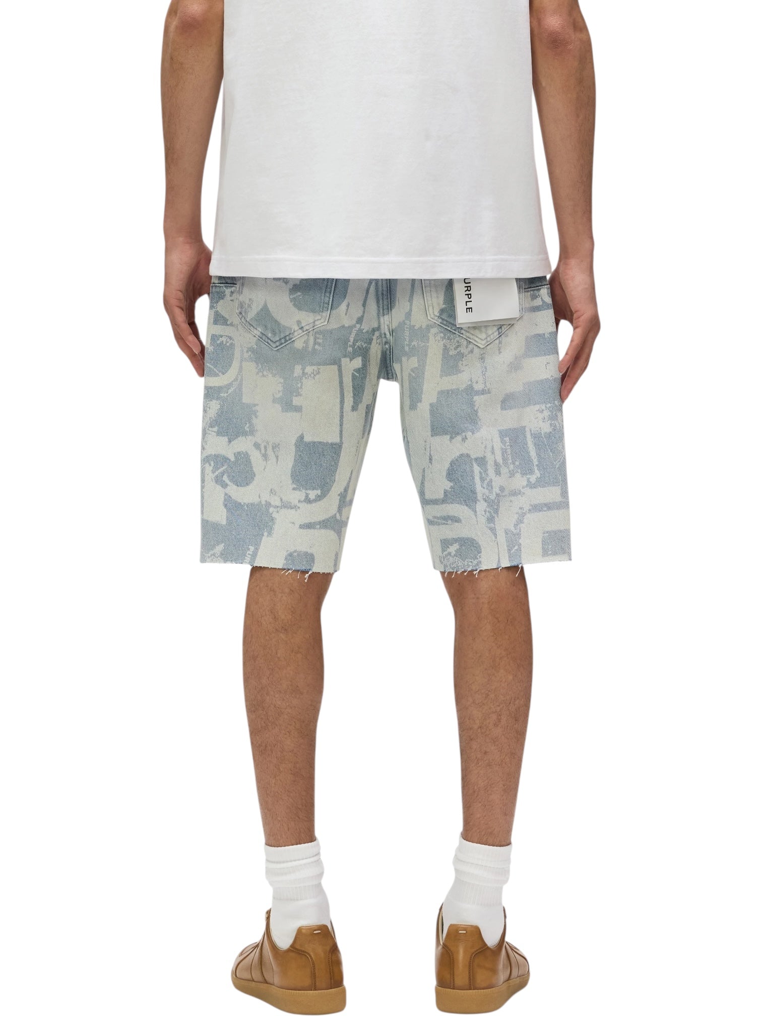 A person is shown from the back wearing a white t-shirt, PURPLE BRAND P059-GRPS325-Grunge Print Indigo Shorts, white socks, tan sneakers, and a visible white tag in one back pocket.