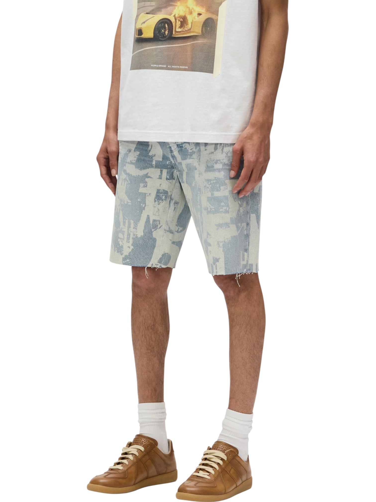 Only the lower half of a person is visible, wearing a printed white t-shirt, PURPLE BRAND P059-GRPS325-Grunge Print Shorts in Indigo, white socks, and brown sneakers.