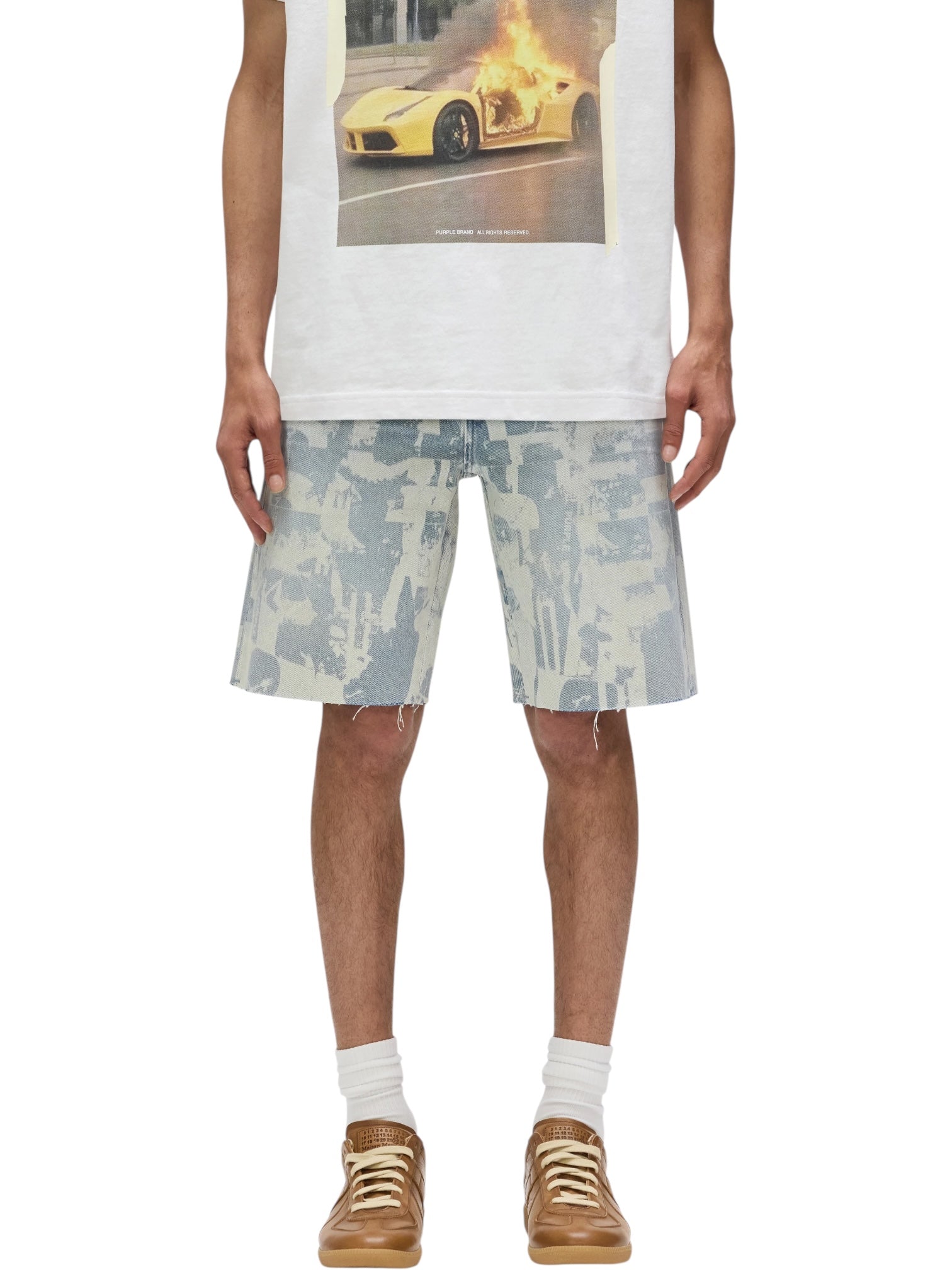 A person wearing a white t-shirt with a car graphic, PURPLE BRAND P059-GRPS325-Grunge Print Shorts in Indigo, white socks, and brown sneakers, standing against a white background.