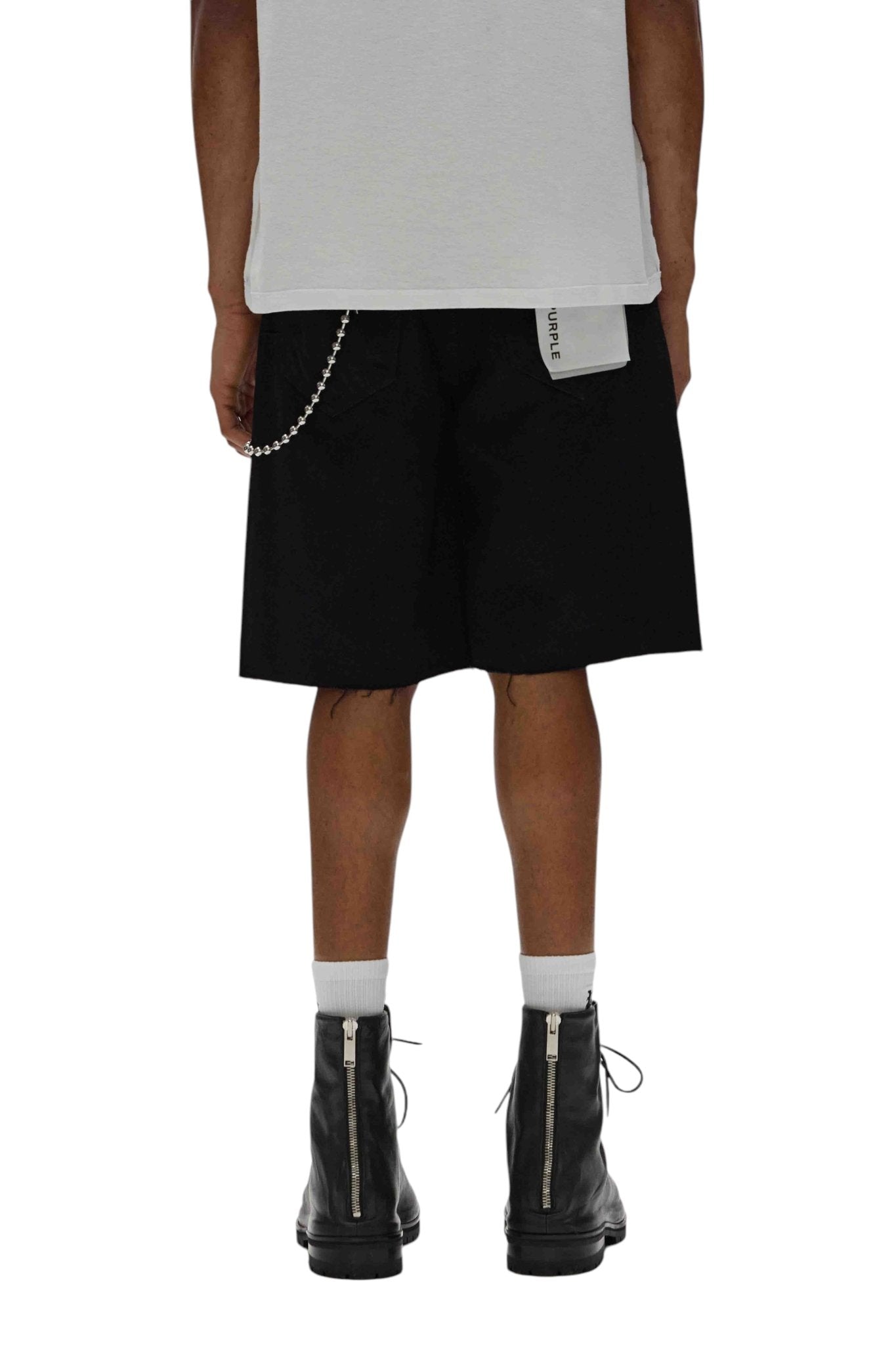 A person in a relaxed fit white T-shirt and PURPLE BRAND's P059-MDSB Midnight Shorts in black, which feature a side chain, stands in black boots with visible zippers, facing away.