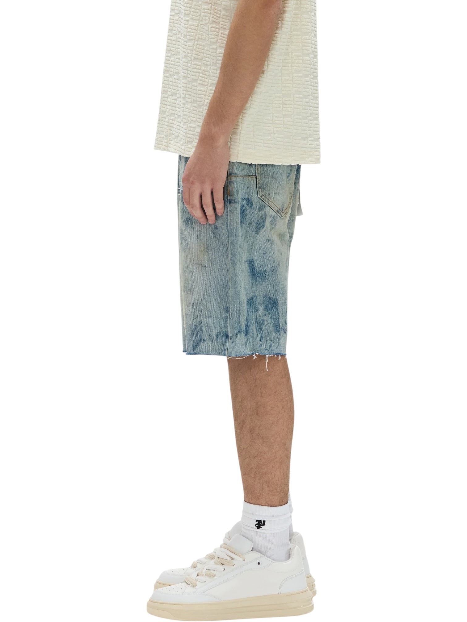 A person standing sideways wears a textured white shirt, PURPLE BRAND P059-MXPS Mixed Paint Shorts Indigo, white crew socks, and white sneakers against a white background.