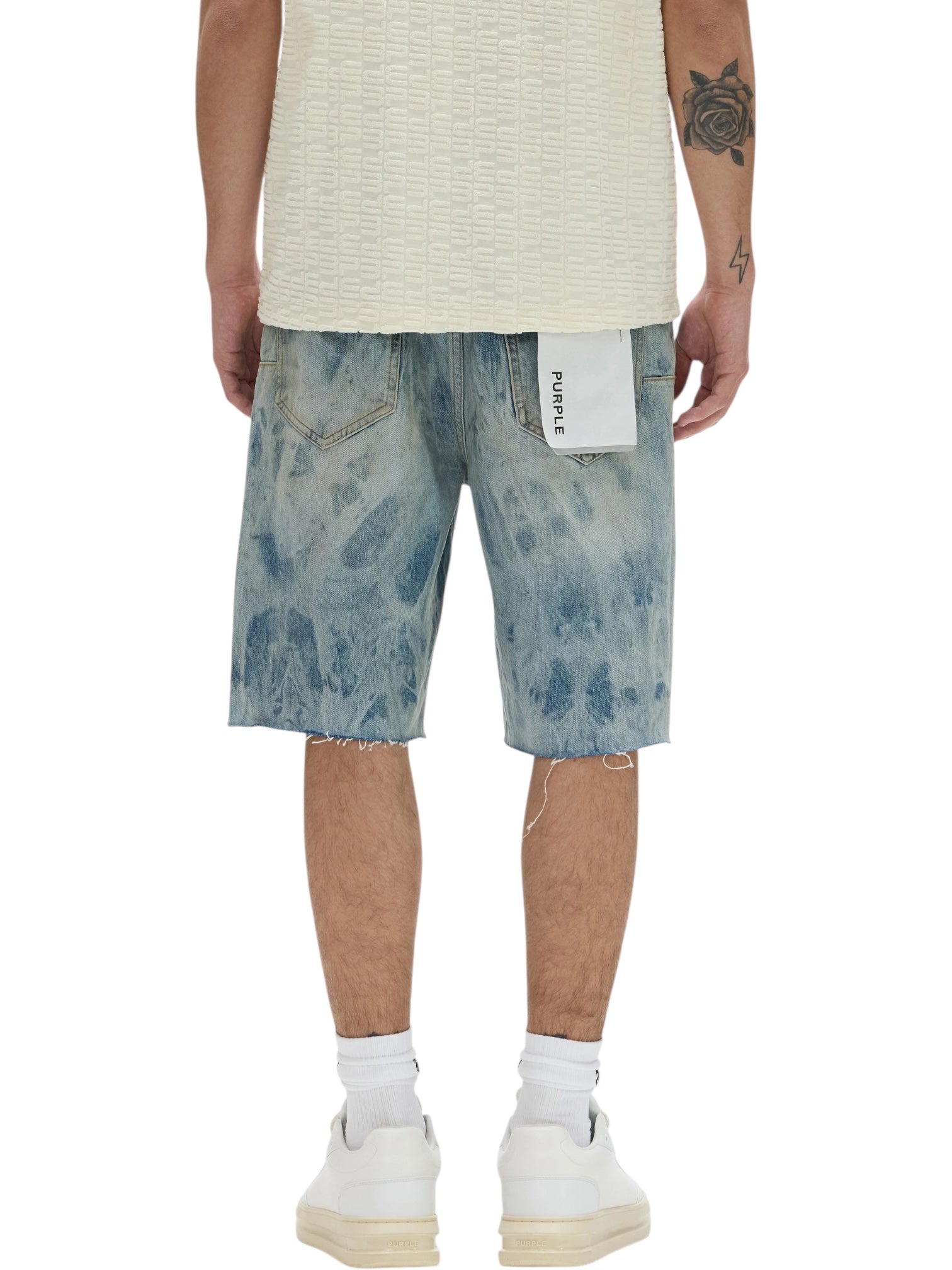 A person is shown from the back wearing a textured white shirt and PURPLE BRAND P059-MXPS Mixed Paint Shorts Indigo, featuring light blue denim, relaxed fit, frayed hems, and a white tag on the pocket. White socks and sneakers complete the look.