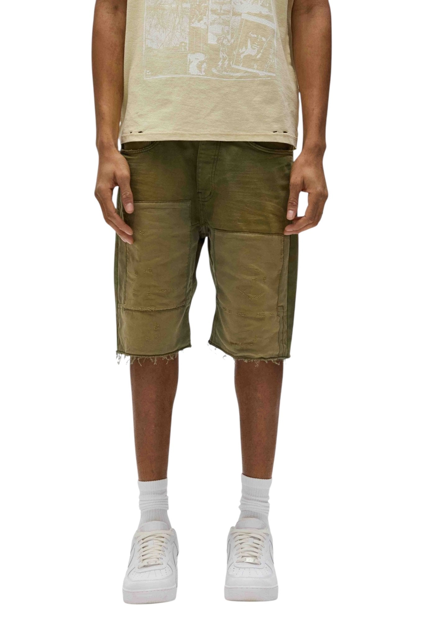 A person wearing a beige t-shirt, the Purple Brand P059-PGSO Patchwork Short Overdye Grn with 5-pocket styling, and white sneakers stands with arms by their sides against a plain background.