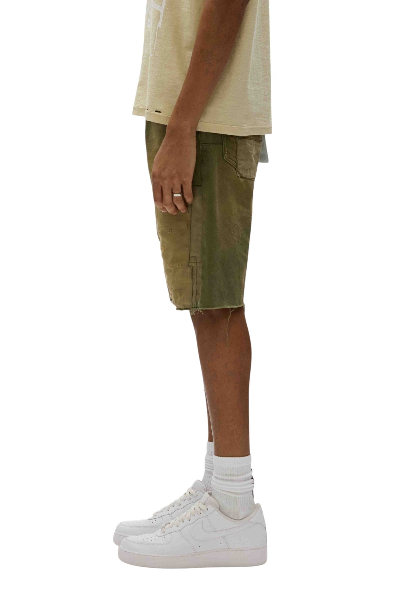 A person in a light shirt and PURPLE BRAND's Patchwork Short Overdye Grn, featuring a relaxed fit and two-tone green design with 5-pocket styling, stands in profile against a plain backdrop, complemented by white socks and crisp white sneakers.