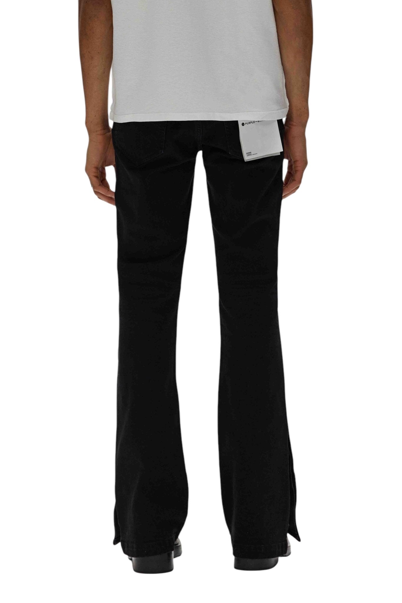 A person is seen from the back wearing a white shirt and black flared pants featuring a visible label tag, similar to PURPLE BRAND's P067-FCNB Flare Crystal Side Snap Blk jeans.