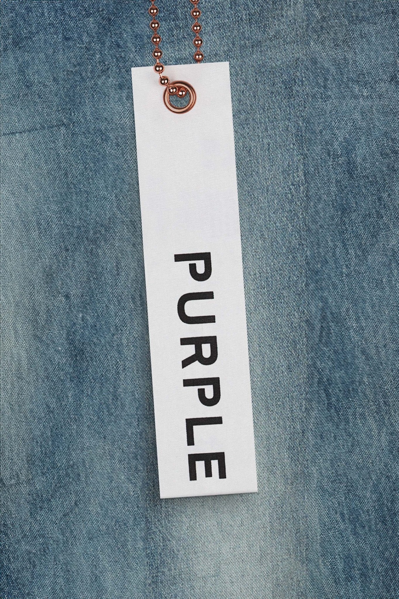 A white tag with "PURPLE BRAND" in bold black letters hangs from a copper-colored chain against a blue fabric background, evocative of the Purple Brand P072-SSPW Super Stack Flare Mid Indigo.