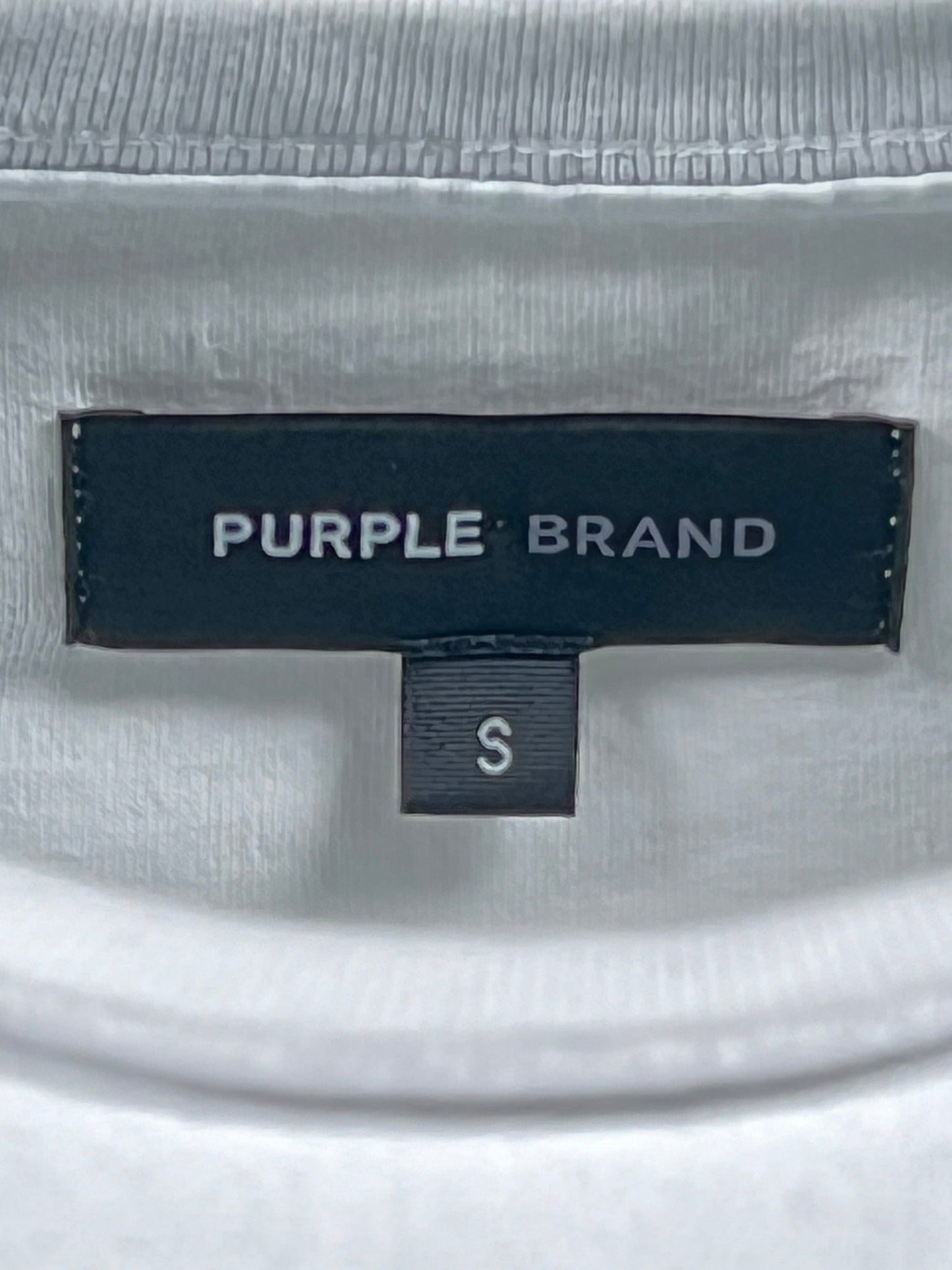 Close-up image of a clothing label with the text "PURPLE BRAND P101-JDBW TEXTURED NSIDE OUT TEE WHITE" in white on a dark tag and a smaller tag below it marked with size "S".