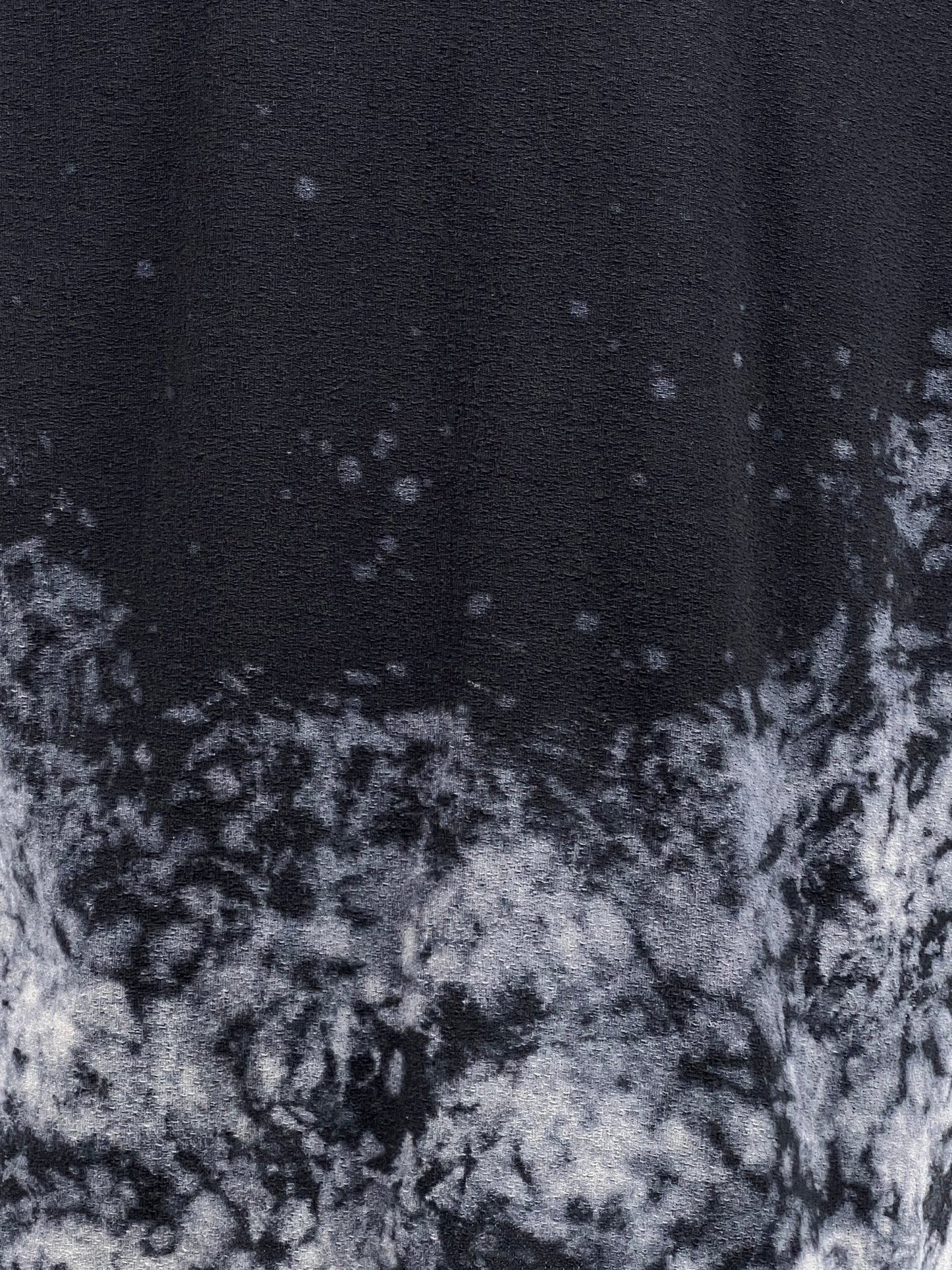 A close-up of the PURPLE BRAND P101-JWBB TEXTURED INSIDE OUT TEE BLACK featuring a white tie-dye pattern near the bottom. The texture appears soft, with an irregular distribution of dye creating a stippled effect. The 100% cotton material adds to its comfort and quality.