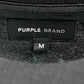 Close-up of a clothing label reading "PURPLE BRAND," featuring a gothic Purple Brand logo and size tag "M" underneath. The garment, identified as the Purple Brand P104-JGBB Textured Jersey Tee Black, is crafted from 100% cotton and presents itself as a premium black jersey with a dark and textured design.
