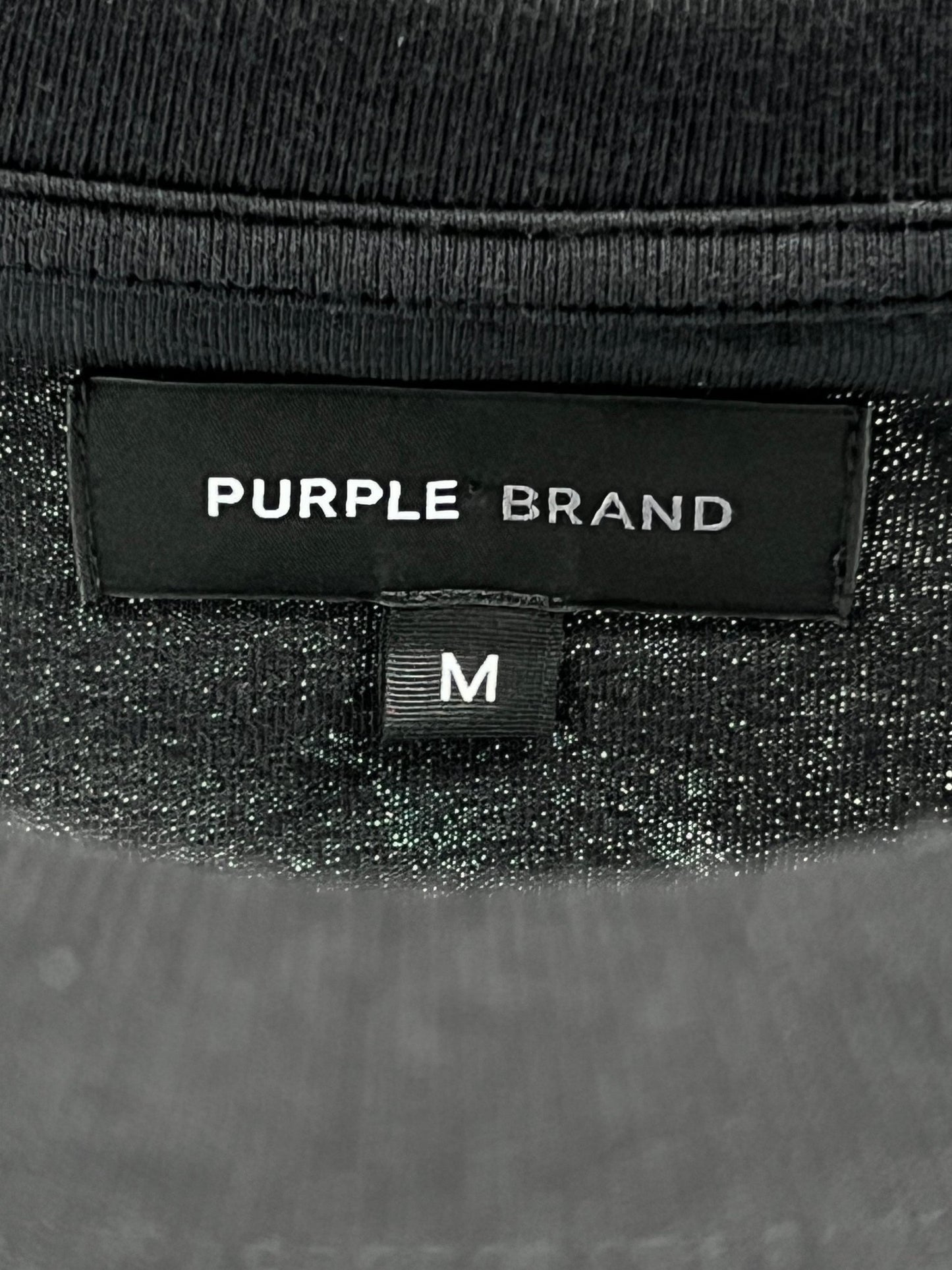 Close-up of a clothing label reading "PURPLE BRAND," featuring a gothic Purple Brand logo and size tag "M" underneath. The garment, identified as the Purple Brand P104-JGBB Textured Jersey Tee Black, is crafted from 100% cotton and presents itself as a premium black jersey with a dark and textured design.