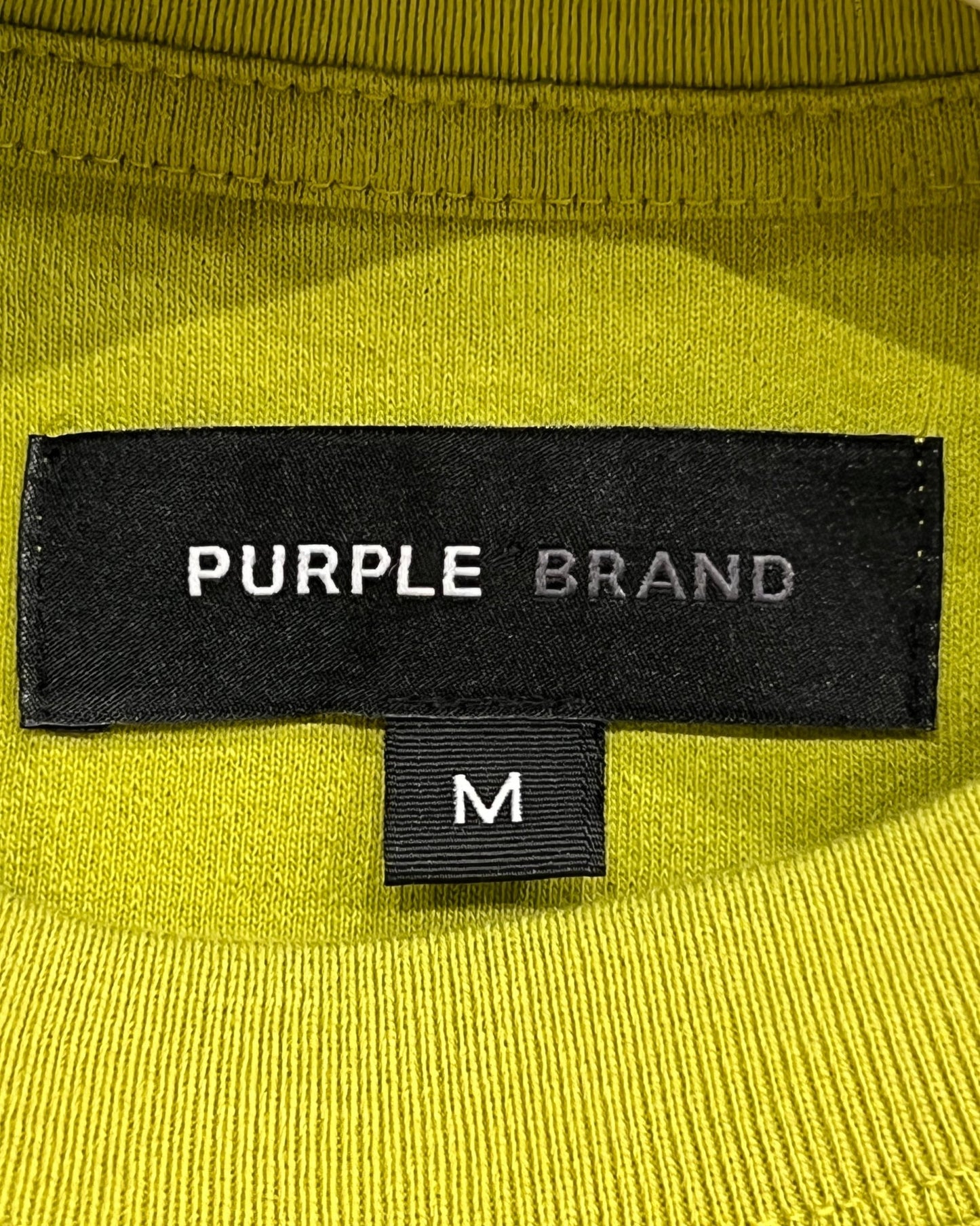 Close-up of a black clothing label reading "PURPLE BRAND" on a bright yellow crewneck tee with a size tag marked 'M'.