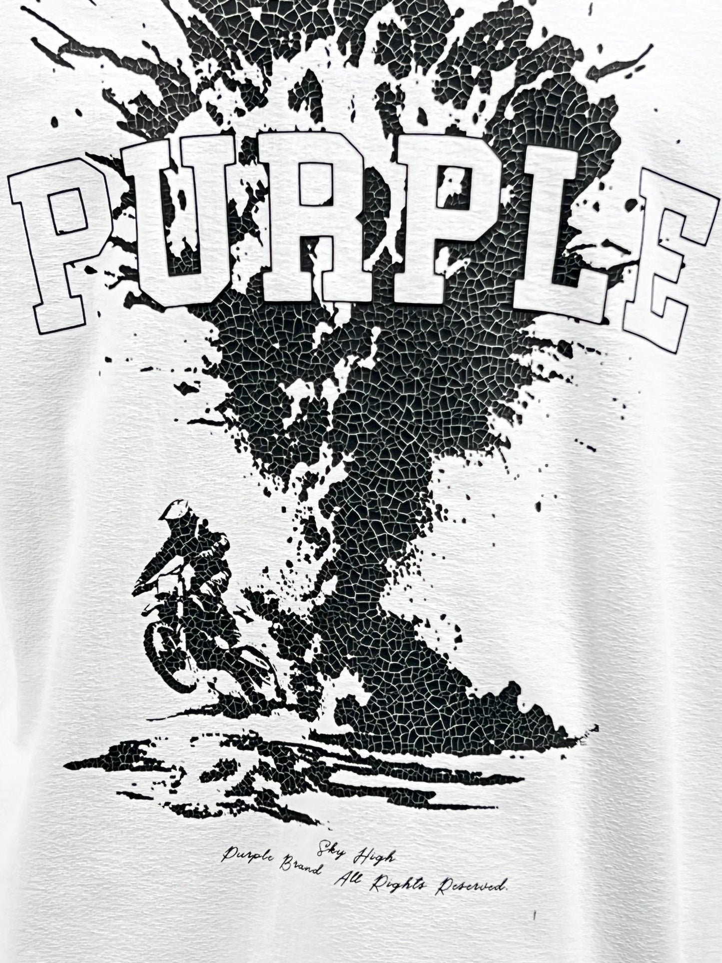 Introducing the Purple Brand P104-JSBW Textured Jersey Tee in White, crafted from 100% cotton to ensure premium quality. This stylish jersey knit shirt features bold cracked-texture "PURPLE" lettering above a striking black graphic of a cyclist surrounded by splashes. The design also includes the phrases "Doyle Brand" and "All Rights Reserved," highlighting its exceptional craftsmanship under the PURPLE BRAND label.