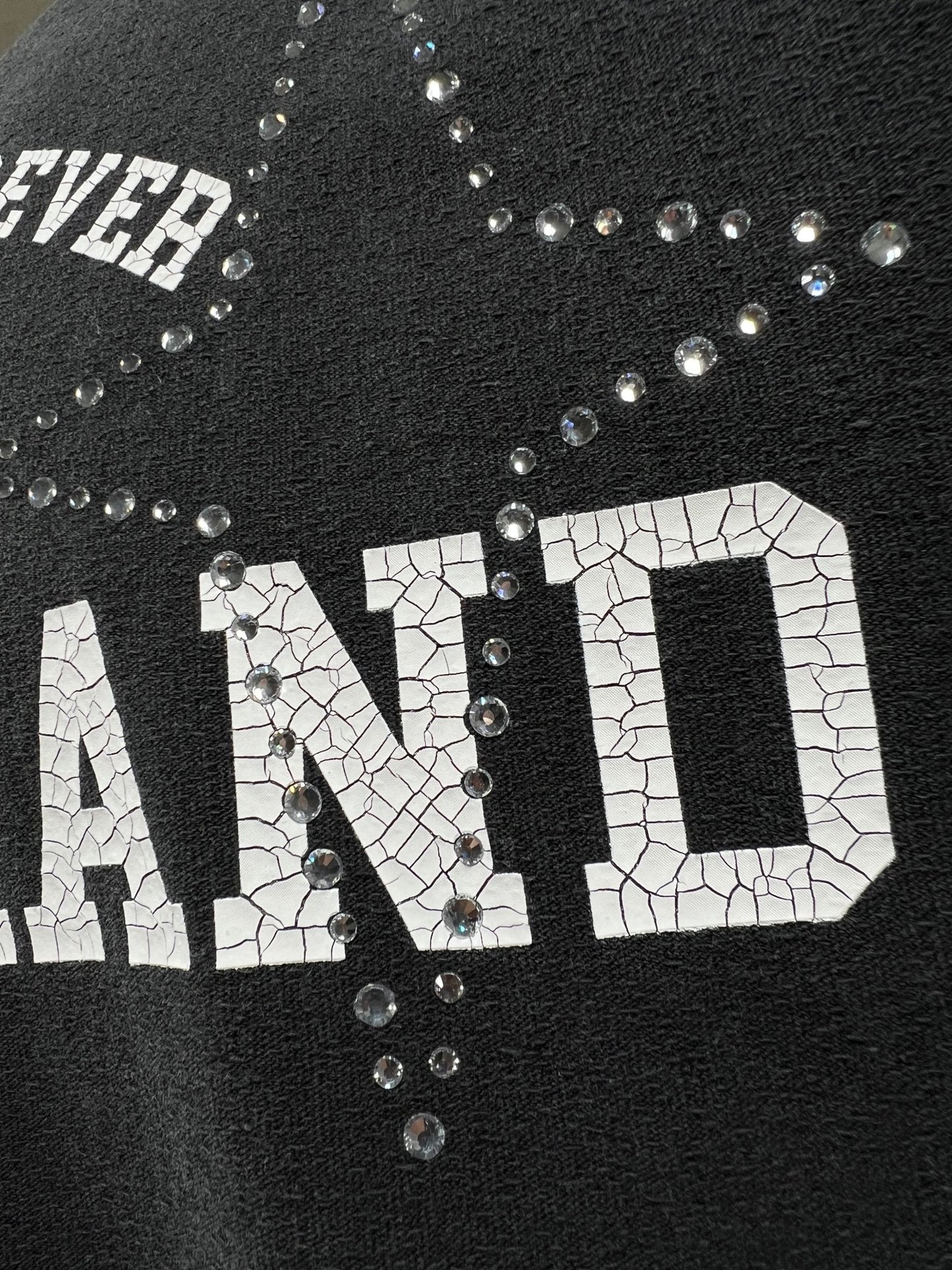 Close-up of a black cotton fabric with white cracked text that reads "LAND," embellished with small, transparent beads arranged in a starburst pattern like crystal stars. "EVER" is visible in the top left corner, adding a stylish touch to this PURPLE BRAND P104-JSCB TEXTURED JERSEY TEE BLACK by PURPLE BRAND.