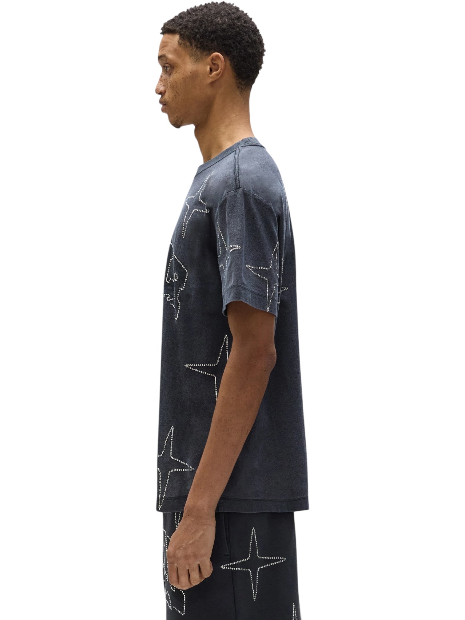A man in profile wears the PURPLE BRAND P104-TBCS325-Textured SS Tee Black Beauty and matching star-patterned shorts with hotfix crystals, set against a white background.