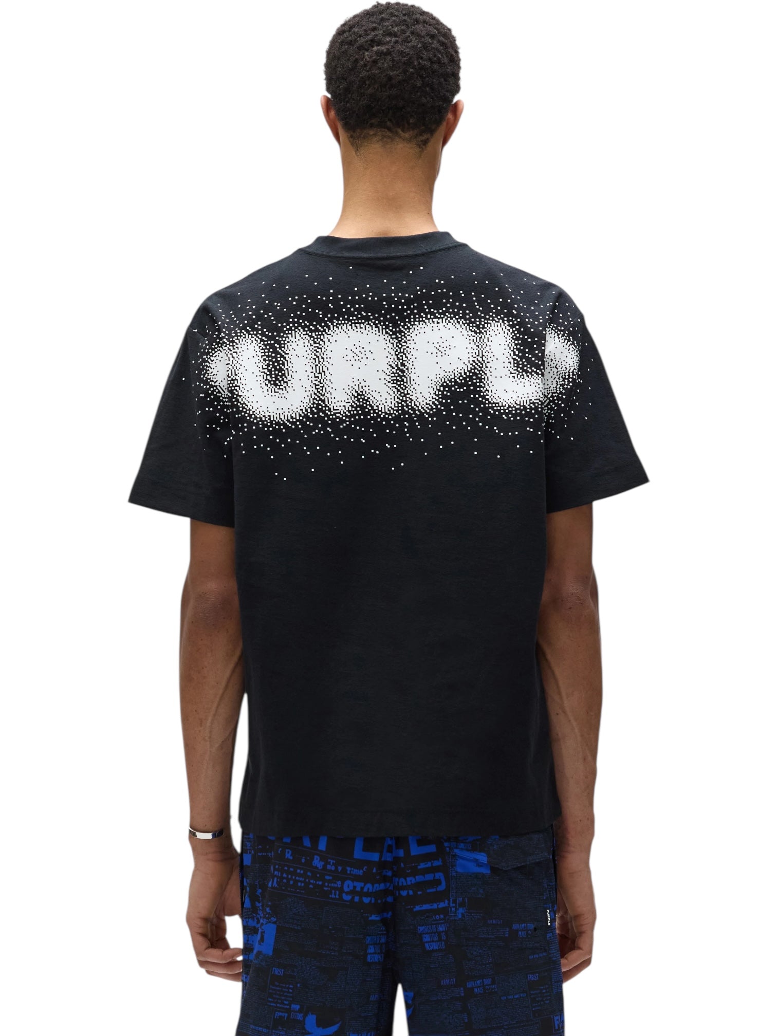 A person is seen from behind, wearing a Purple Brand P104-TBPW Textured Jersey SS tee in black with "PURPLE" in white dotted text, paired with blue graphic shorts.