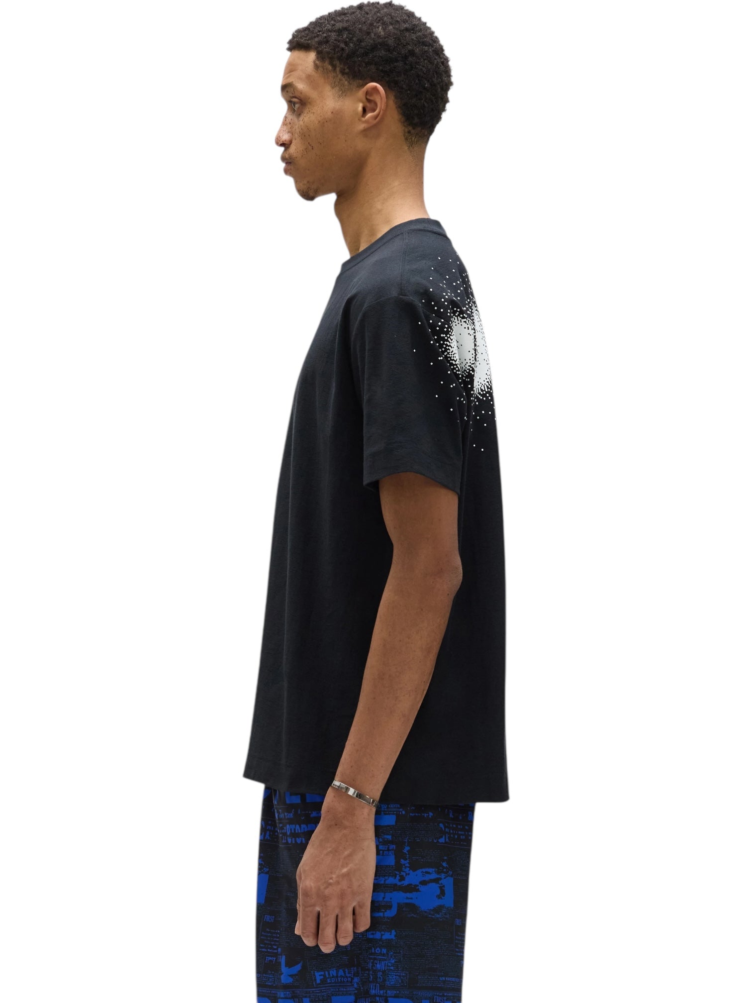 A man in profile wears the PURPLE BRAND P104-TBPW Textured Jersey SS Tee in black, featuring a white splatter design on the back and blue patterned 100% cotton shorts.