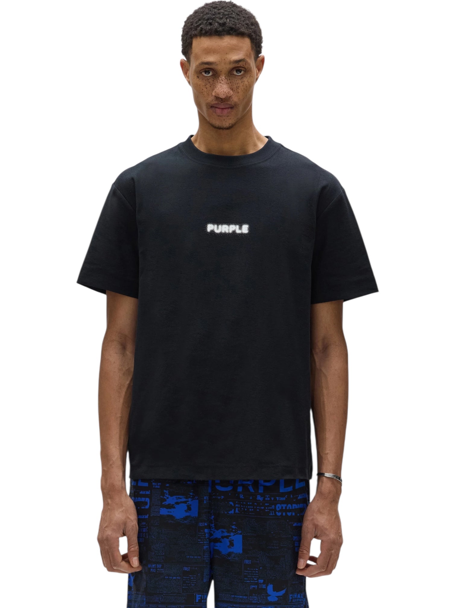 A man stands facing forward wearing a PURPLE BRAND P104-TBPW Textured Jersey SS tee in black with "PURPLE" in white text and blue patterned shorts.