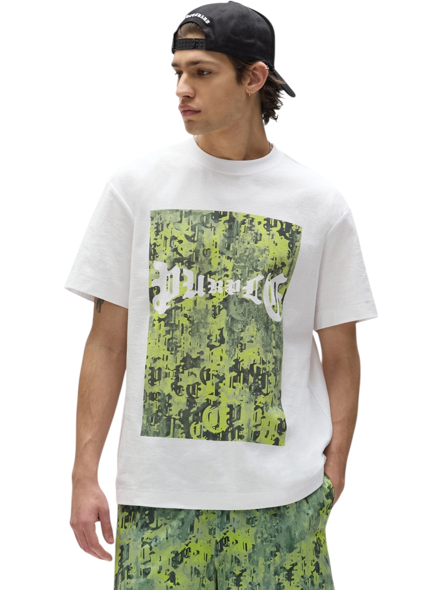 A young man in a backwards black cap and a Purple Brand P104-TWGC325 Textured Jersey SS Tee White, featuring a green and black digital camo print with "MUSARTC," paired with matching green patterned shorts.