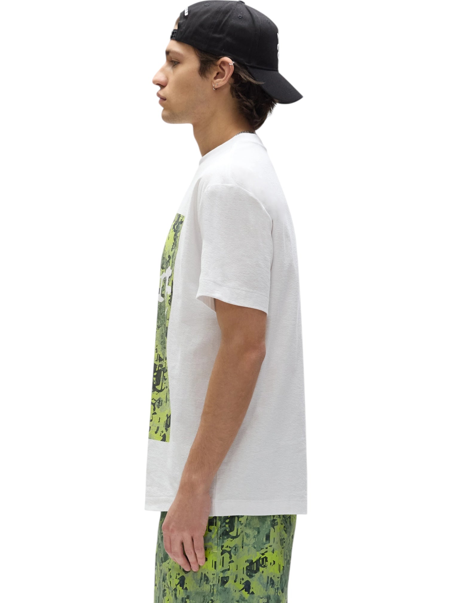 A man in a PURPLE BRAND P104-TWGC325 Textured Jersey SS Tee in white, green digital camo shorts, and a black backwards cap stands in profile against a white background.