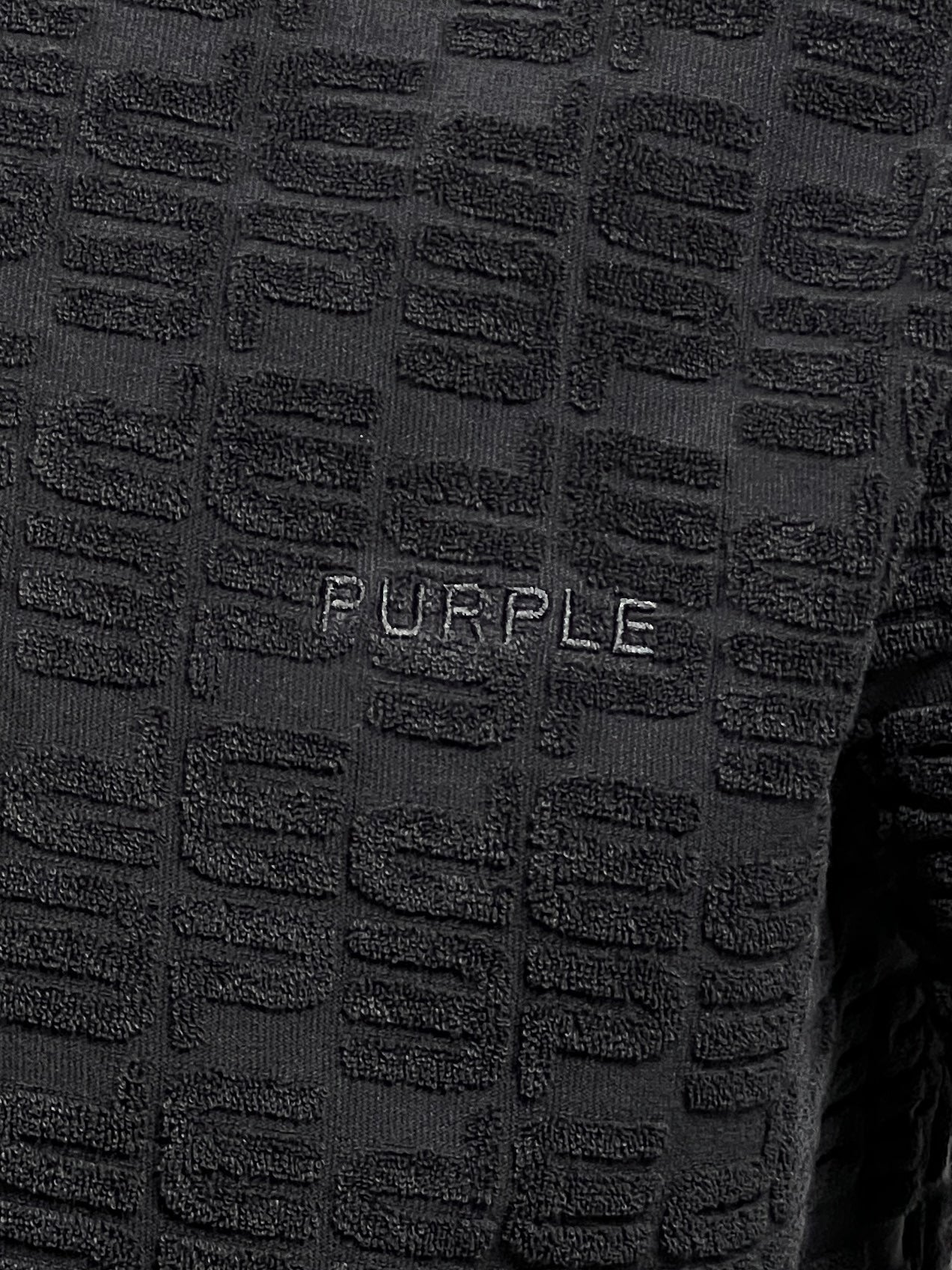 Close-up of a textured black towel-terry fabric with an embossed pattern and the word "PURPLE" embroidered in white, reminiscent of the refined styling found in PURPLE BRAND PURPLE BRAND P138-JTBB TERRY TOWEL POLO BLACK from Pre Fall 2024.