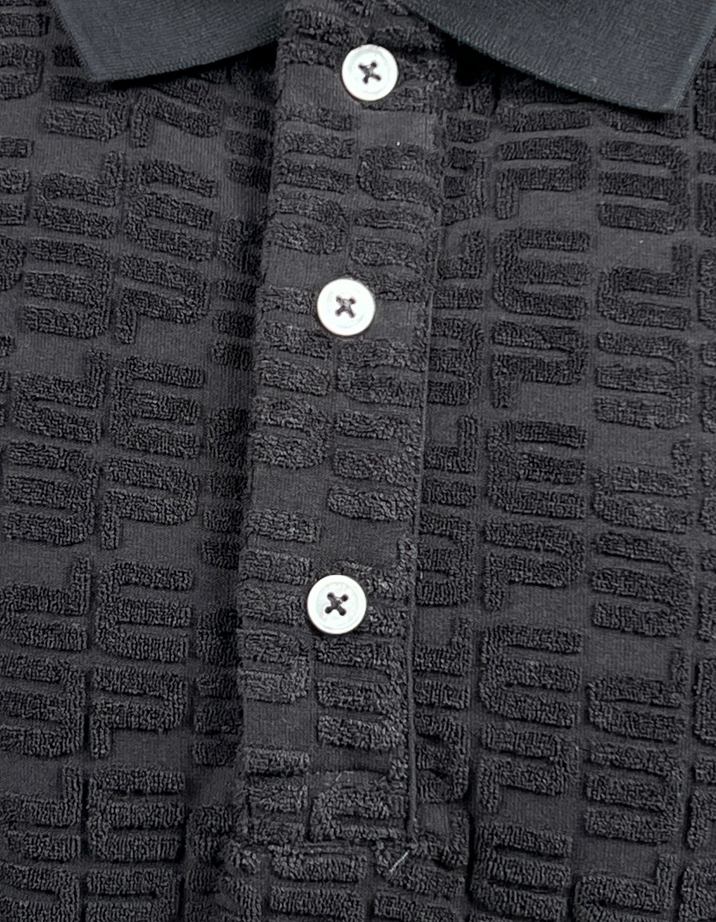 Close-up of a PURPLE BRAND P138-JTBB TERRY TOWEL POLO BLACK made from black towel-terry fabric with a patterned texture and three white buttons on the placket, perfect for Pre Fall 2024.