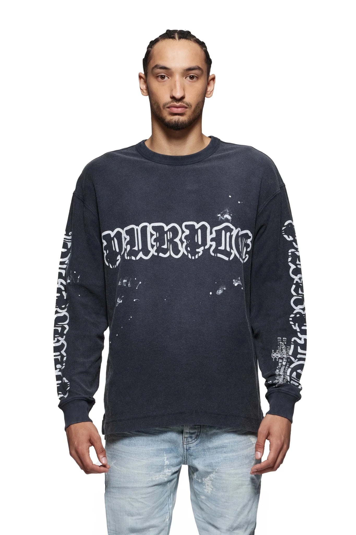 A man wearing the Purple Brand P204-JWGW Textured Jersey LS Tee in black, featuring a gothic logo and white graffiti text on the front and sleeves, paired with light blue jeans against a plain white backdrop.