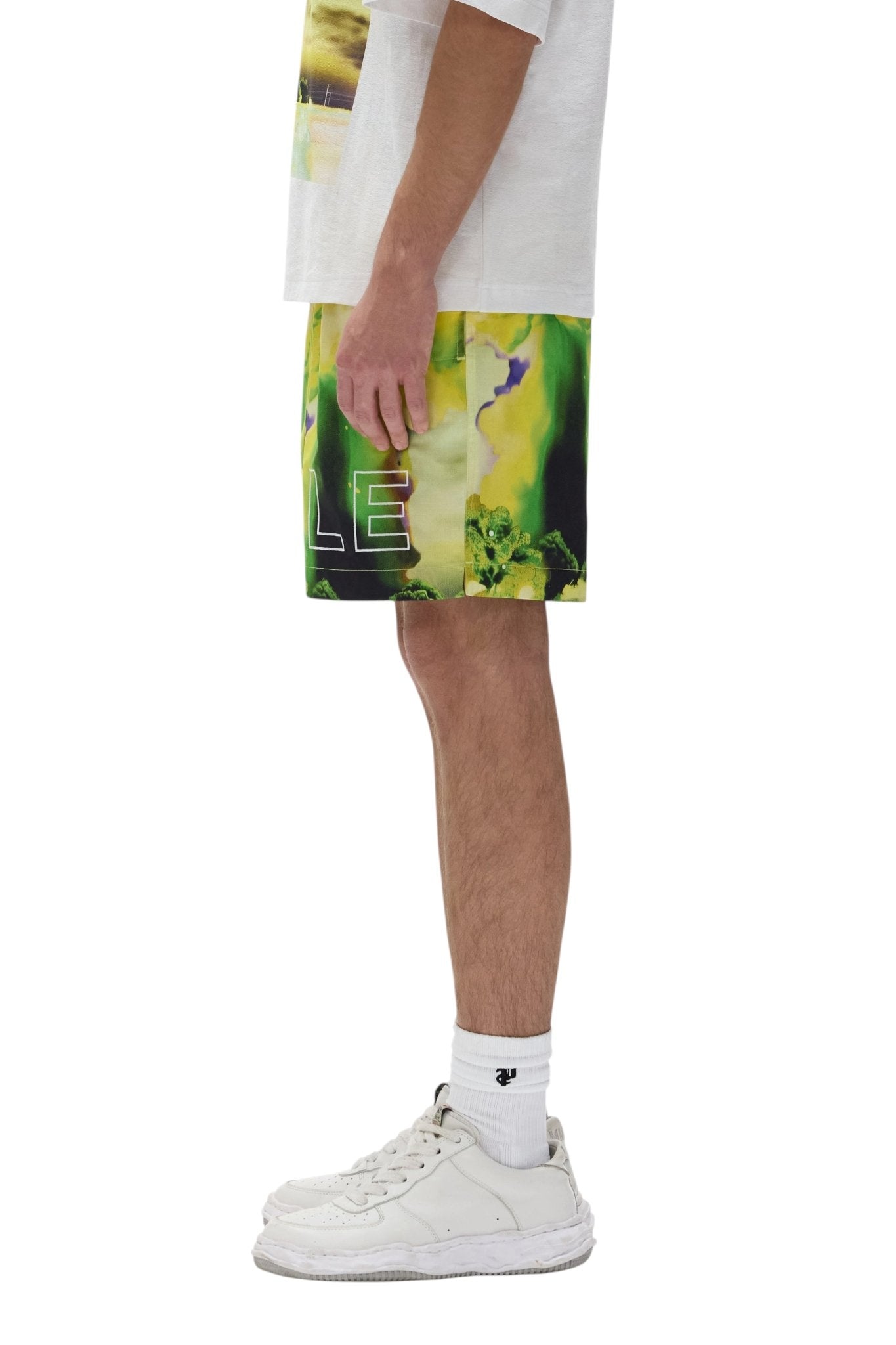 A person stands sideways wearing a white T-shirt, PURPLE BRAND P504-PMFM325-All Round Shorts Yellow with green and yellow patterns, white socks, and white sneakers.