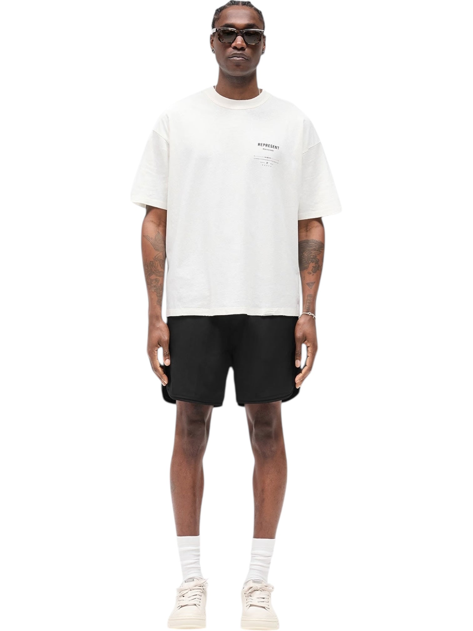 A person wearing sunglasses, black shorts, white socks, white sneakers, and a REPRESENT MLM100286-27 Revere Manor T-shirt in Aged White stands facing forward against a plain white background.