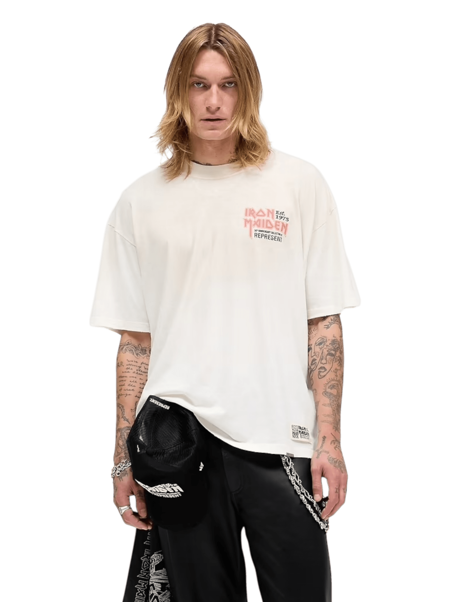 A person with long blond hair wears an oversized REPRESENT Iron Maiden Anniversary T-shirt (Washed Ecru, MLM100579-26), black pants, and holds a black cap in their left hand against a plain background.
