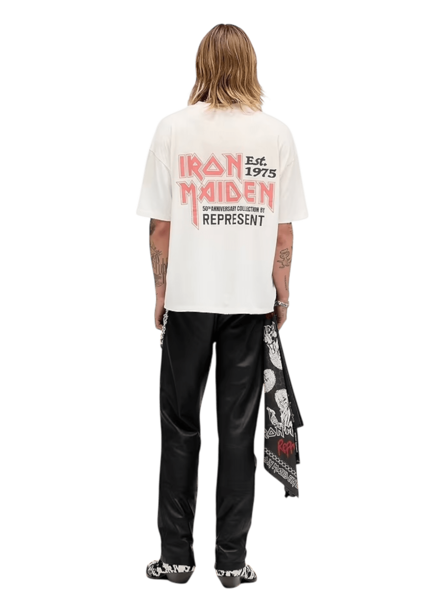 A person with shoulder-length hair faces away in an oversized REPRESENT MLM100579-26 Iron Maiden Anniversary T-shirt Washed Ecru, black pants, printed shoes, and a bandana at the side—perfect vintage rock tee style.