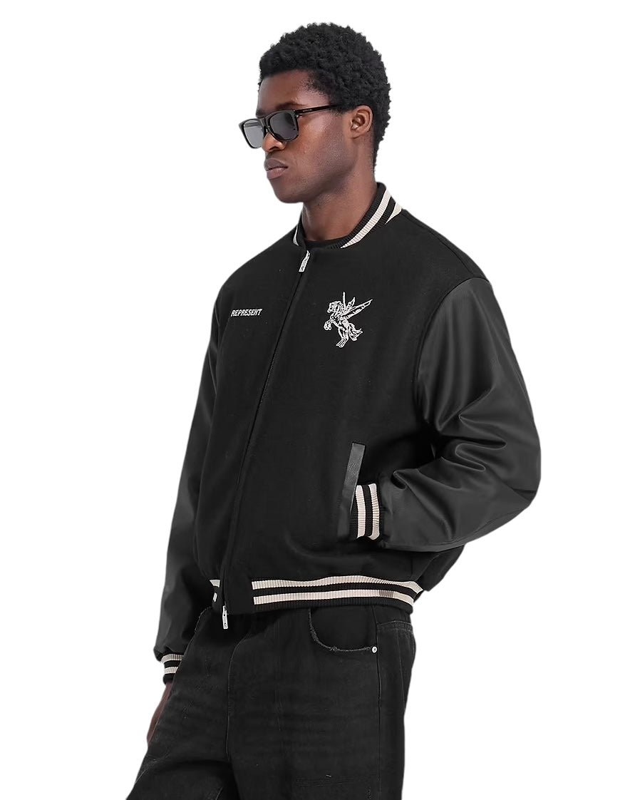 Wearing REPRESENT's sleek Bellerophon Wool Varsity Jacket, featuring RiRi zippers and leather sleeves, a person confidently stands against a white backdrop in chic black sunglasses and matching black pants, exuding an effortlessly cool and modern vibe.