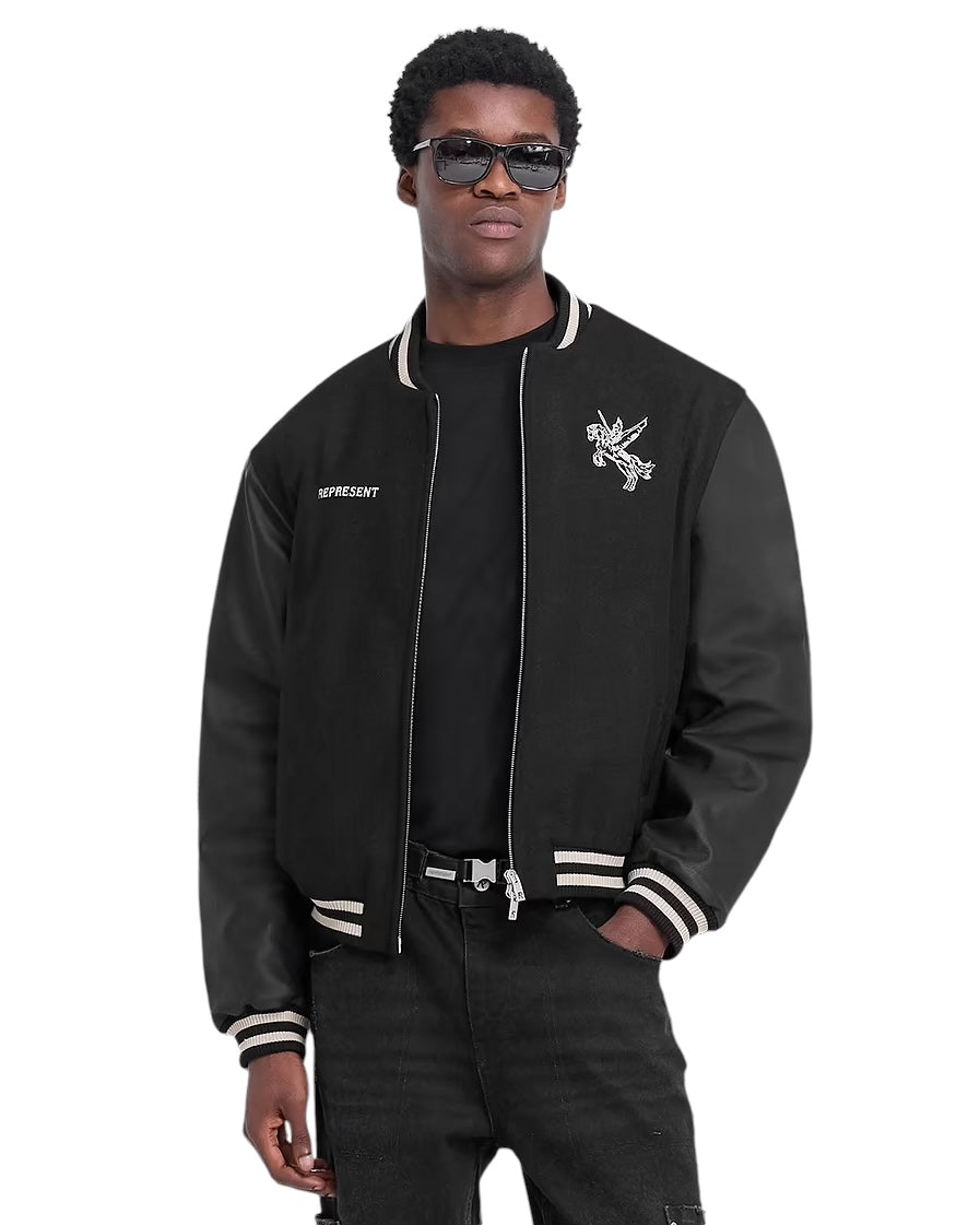 Wearing the REPRESENT MLM1127 Bellerophon Wool Varsity Jacket in black, featuring embroidered mascot designs and RiRi zippers, they exude confidence with sunglasses, one hand casually in a pocket. The look is completed with a sleek black shirt and pants.