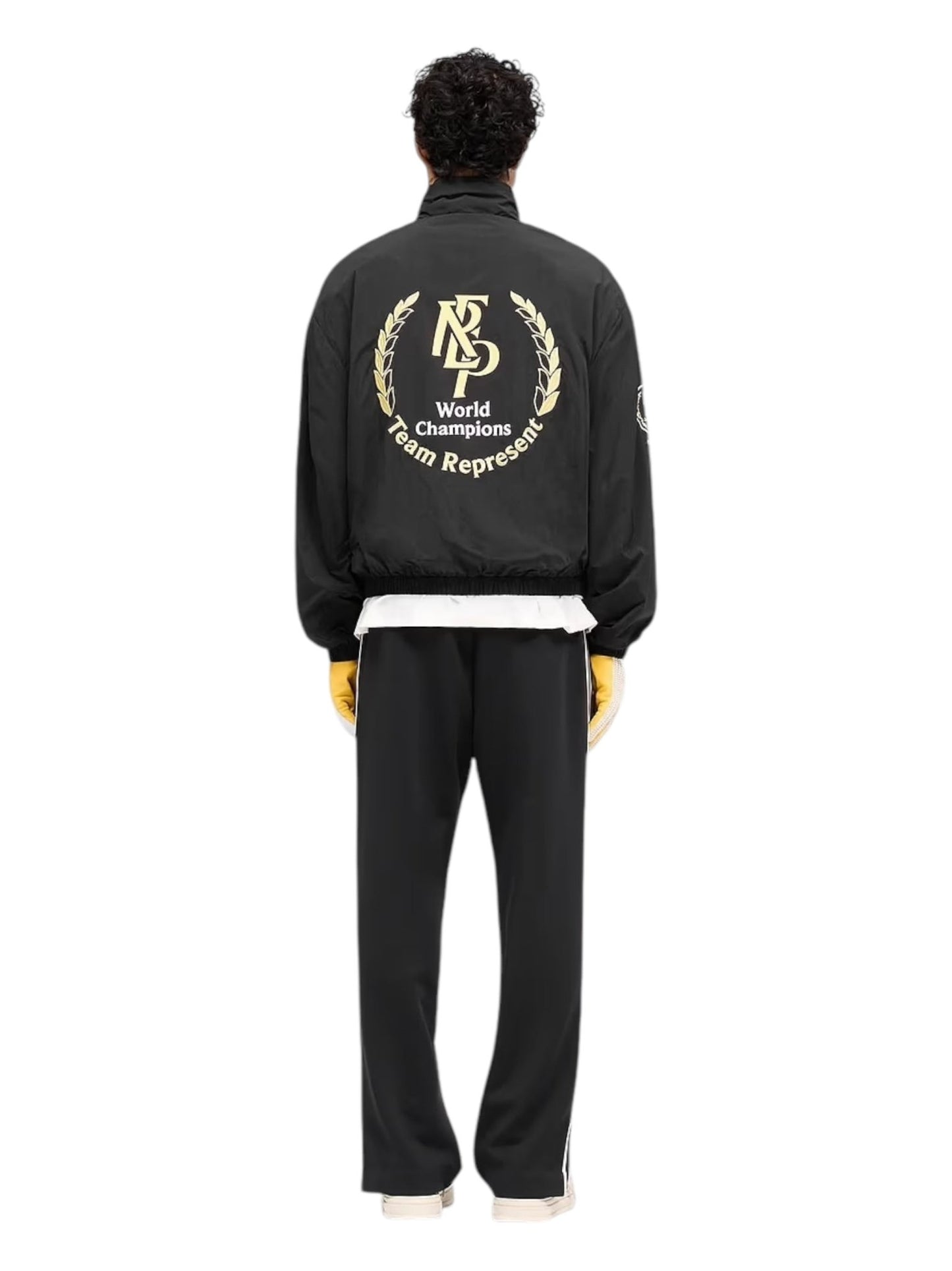 A person wearing the REPRESENT MLM1795 World Championship Jacket in black features a gold laurel design and "World Champions Team Represent" text on the back, paired with black pants and yellow gloves, creating a bold statement.