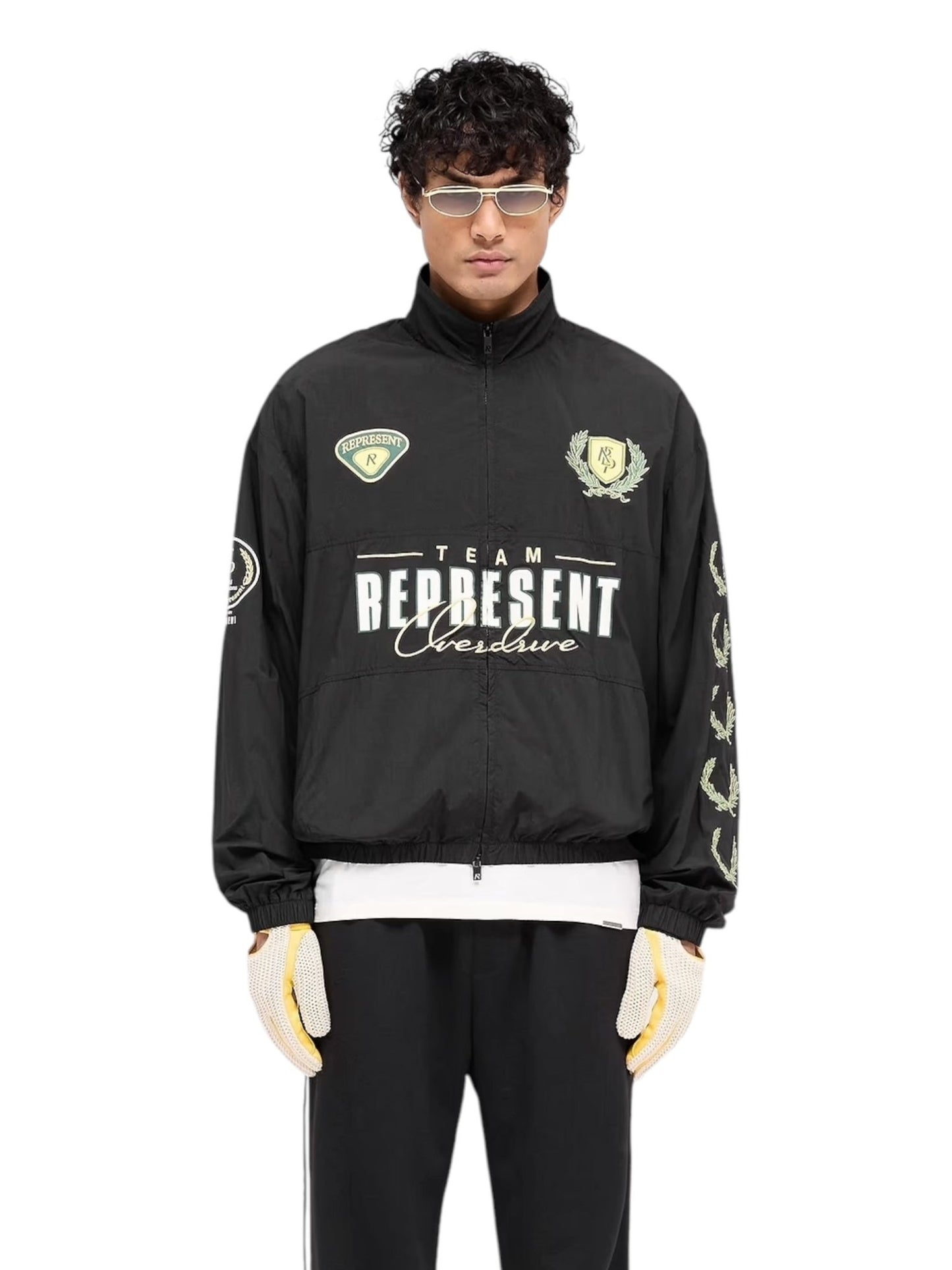 A person confidently stands against a plain background wearing the REPRESENT MLM1795 World Championship Jacket in black with "Team Represent," paired with white gloves, white pants, and sunglasses, embodying a redefined cool style.