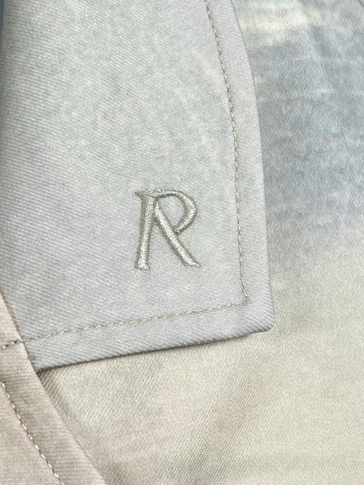 Close-up of a gray fabric with a stitched "R" monogram on the edge of a fold or flap, embodying the elegance of our REPRESENT MLM206-137 HIGHER TRUTH PRINTED SHIRT MULT with its luxury boxy fit.