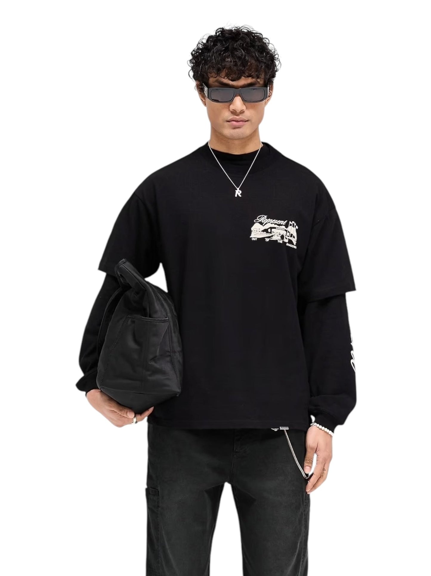 A person in a Represent MLM41063 Raceway Tee in Jet Black, dark pants, and sunglasses holds a black bag against a white background.