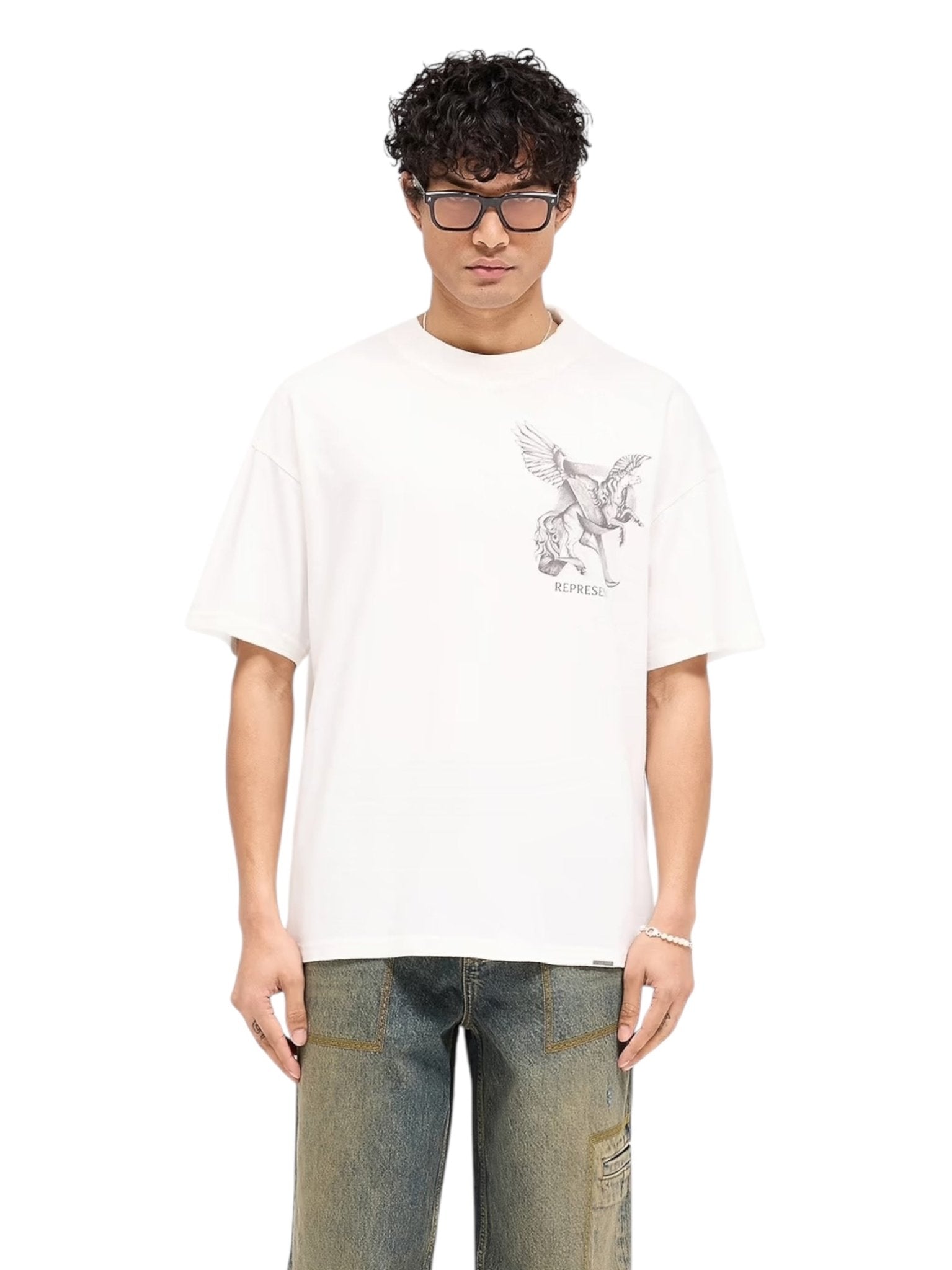 A person wearing glasses and the REPRESENT MLM41066 Elegance in Motion Tee in white, crafted from soft jersey cotton with a winged horse graphic, stands stylishly against a plain background, complemented by classic blue jeans.