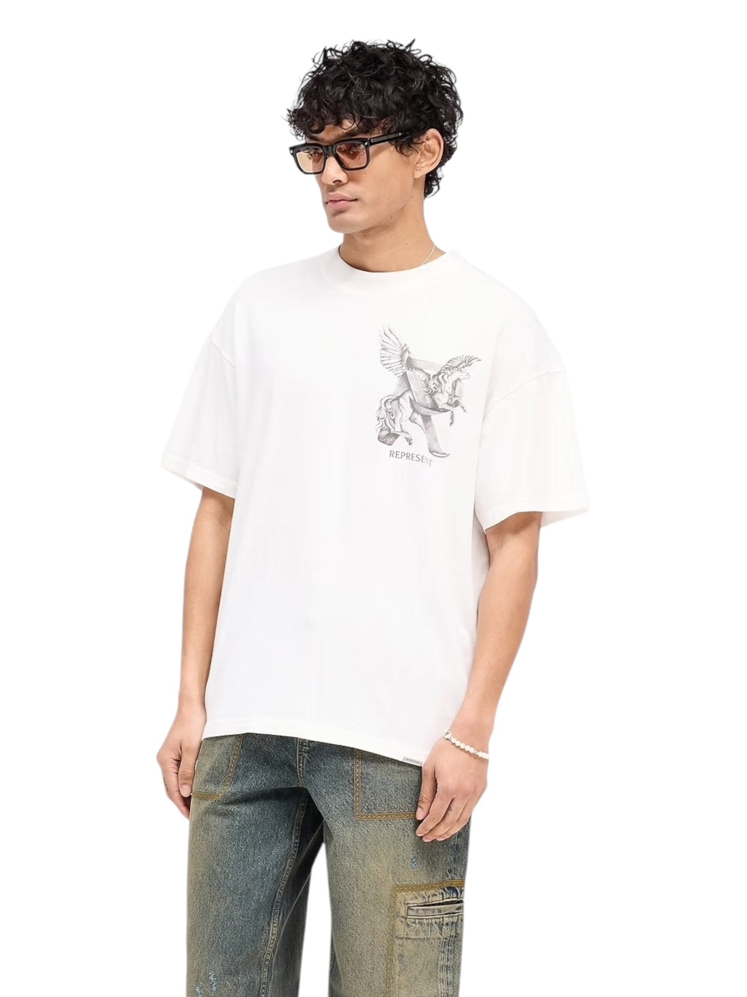 Someone in sunglasses, an oversized Represent MLM41066 Elegance in Motion Tee in White by REPRESENT, made of jersey cotton with a Pegasus design, paired with blue jeans featuring pocket detail, stands against a plain background.