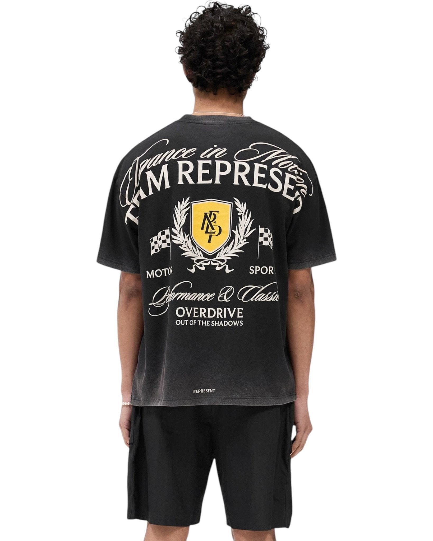 Individual dressed in a REPRESENT MLM41069 Crest T-shirt in vintage black, featuring back graphics, paired with black shorts, facing away.