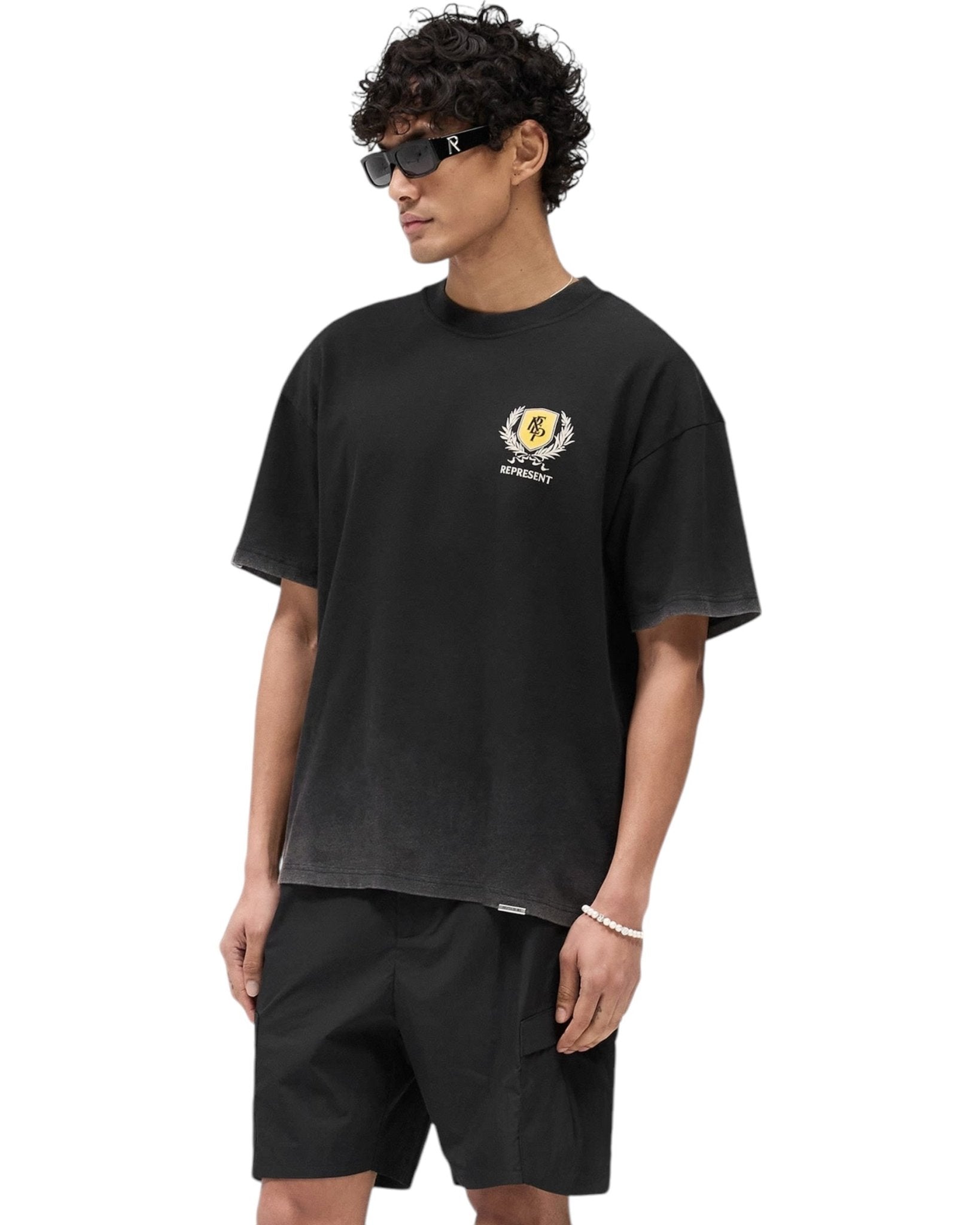 A person in a REPRESENT MLM41069 Crest T-shirt Vintage Black and black shorts, accessorized with sunglasses and a bracelet, stands against a plain background.