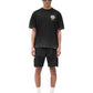 A person stands wearing a Represent MLM41069 Crest T-shirt in Vintage Black with the REPRESENT logo, black shorts, black sneakers, white socks, and dark sunglasses.
