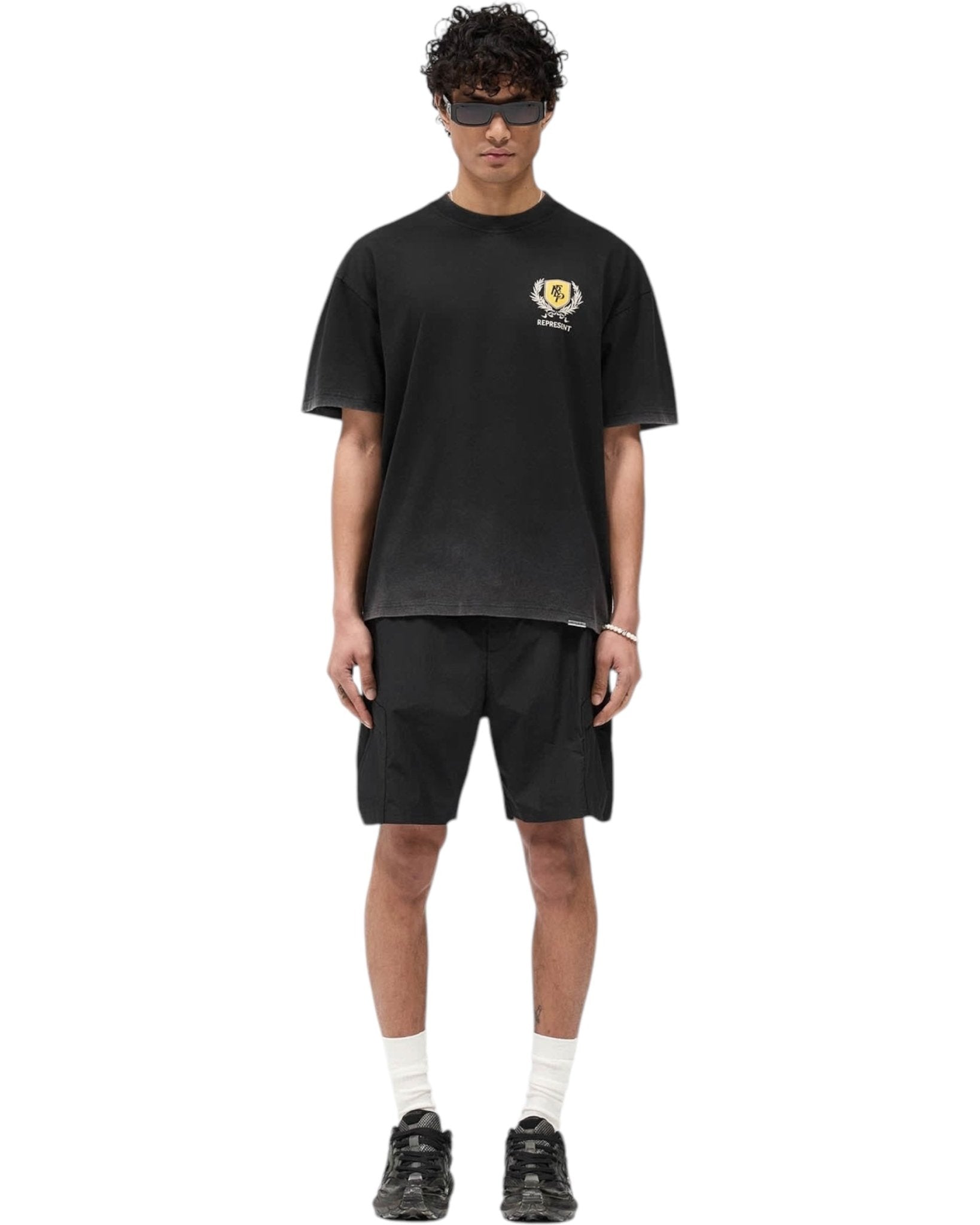 A person stands wearing a Represent MLM41069 Crest T-shirt in Vintage Black with the REPRESENT logo, black shorts, black sneakers, white socks, and dark sunglasses.