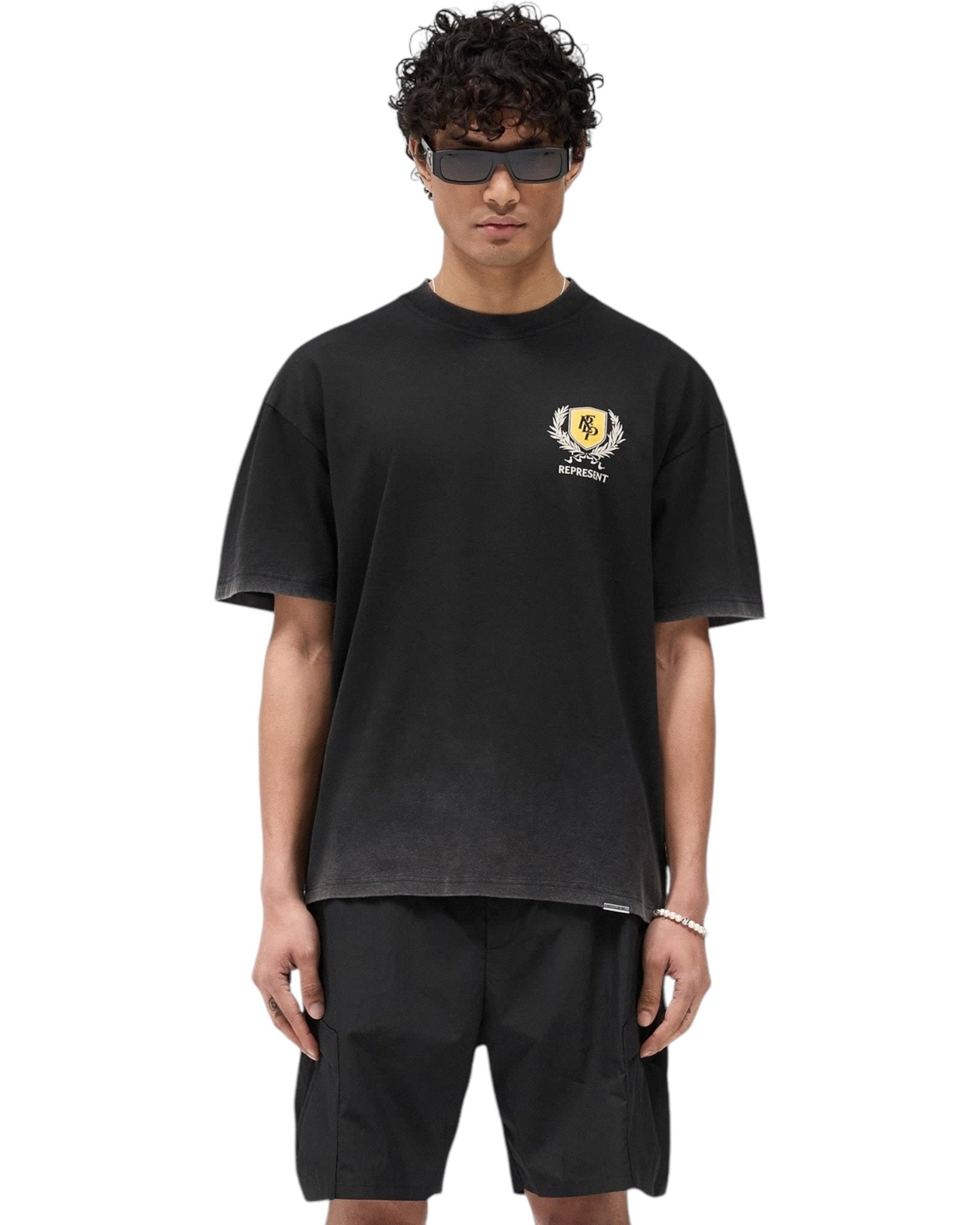 A person wearing a Represent MLM41069 Crest T-shirt in vintage black, paired with black shorts and sunglasses, stands against a white background.
