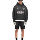 Someone is wearing a vintage-style Represent MLM41144 World Championship Hoodie in black with black shorts. The outfit is finished with a beige cap, sunglasses, gloves, and black socks paired with white sneakers against a crisp white background.