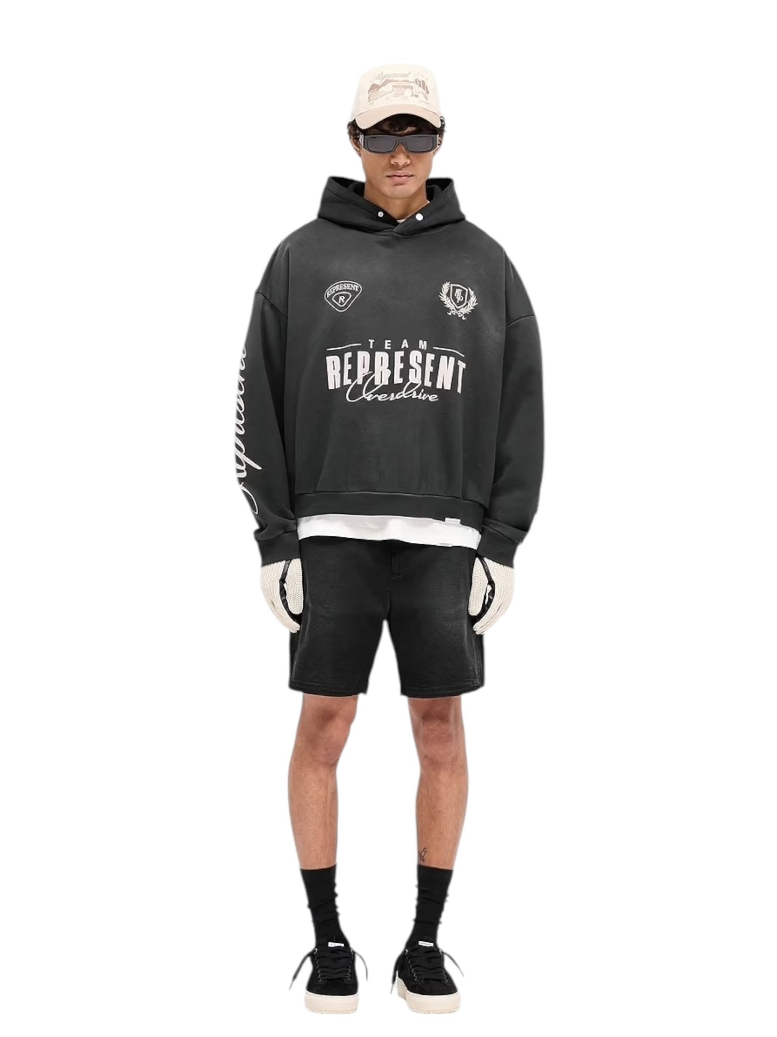 Someone is wearing a vintage-style Represent MLM41144 World Championship Hoodie in black with black shorts. The outfit is finished with a beige cap, sunglasses, gloves, and black socks paired with white sneakers against a crisp white background.