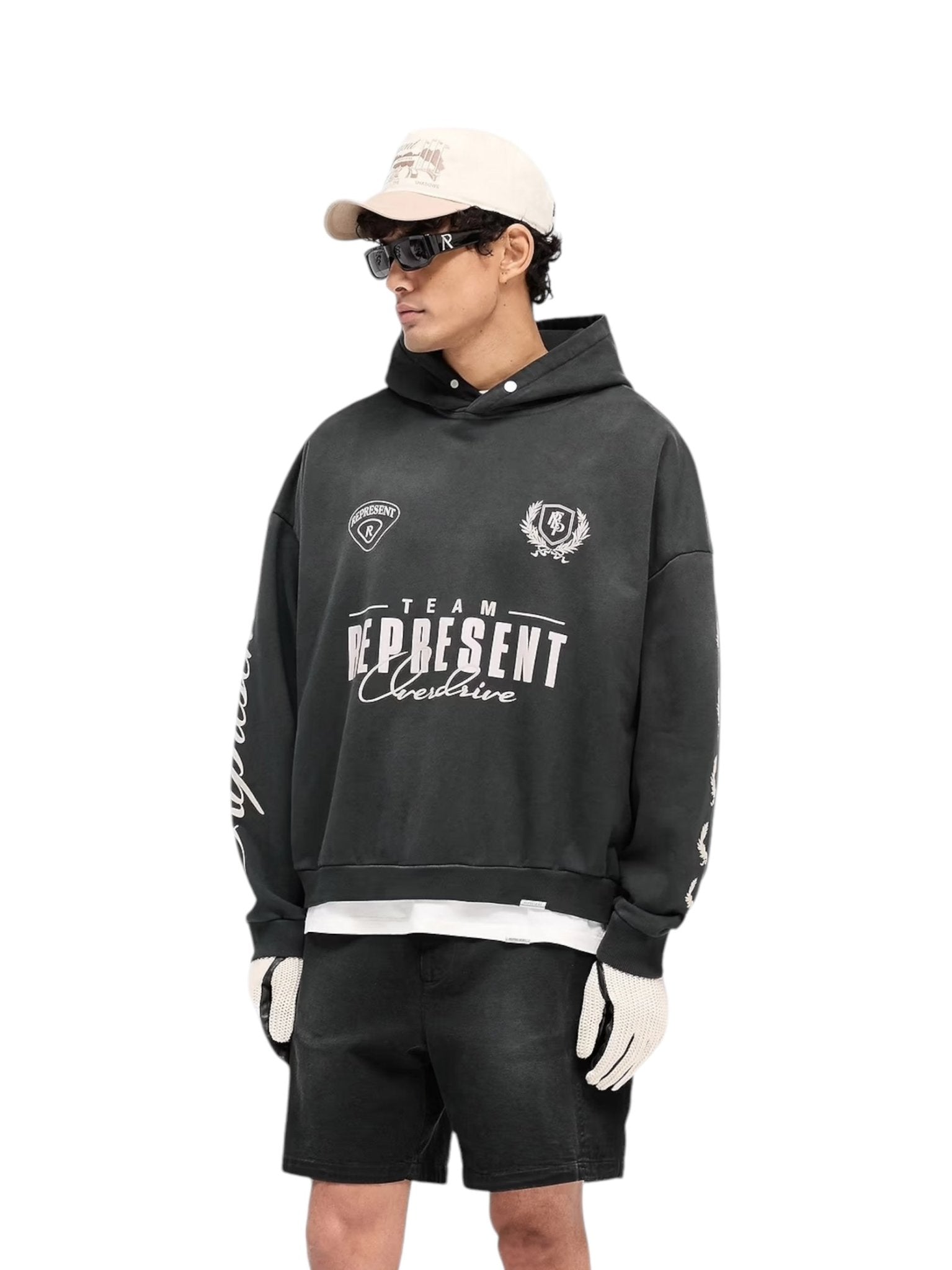 A person exudes confidence in a vintage style, donning the oversized Represent MLM41144 World Championship Hoodie in Black with white text. They pair it with black shorts, white gloves, sunglasses, and a cap against a simple backdrop.