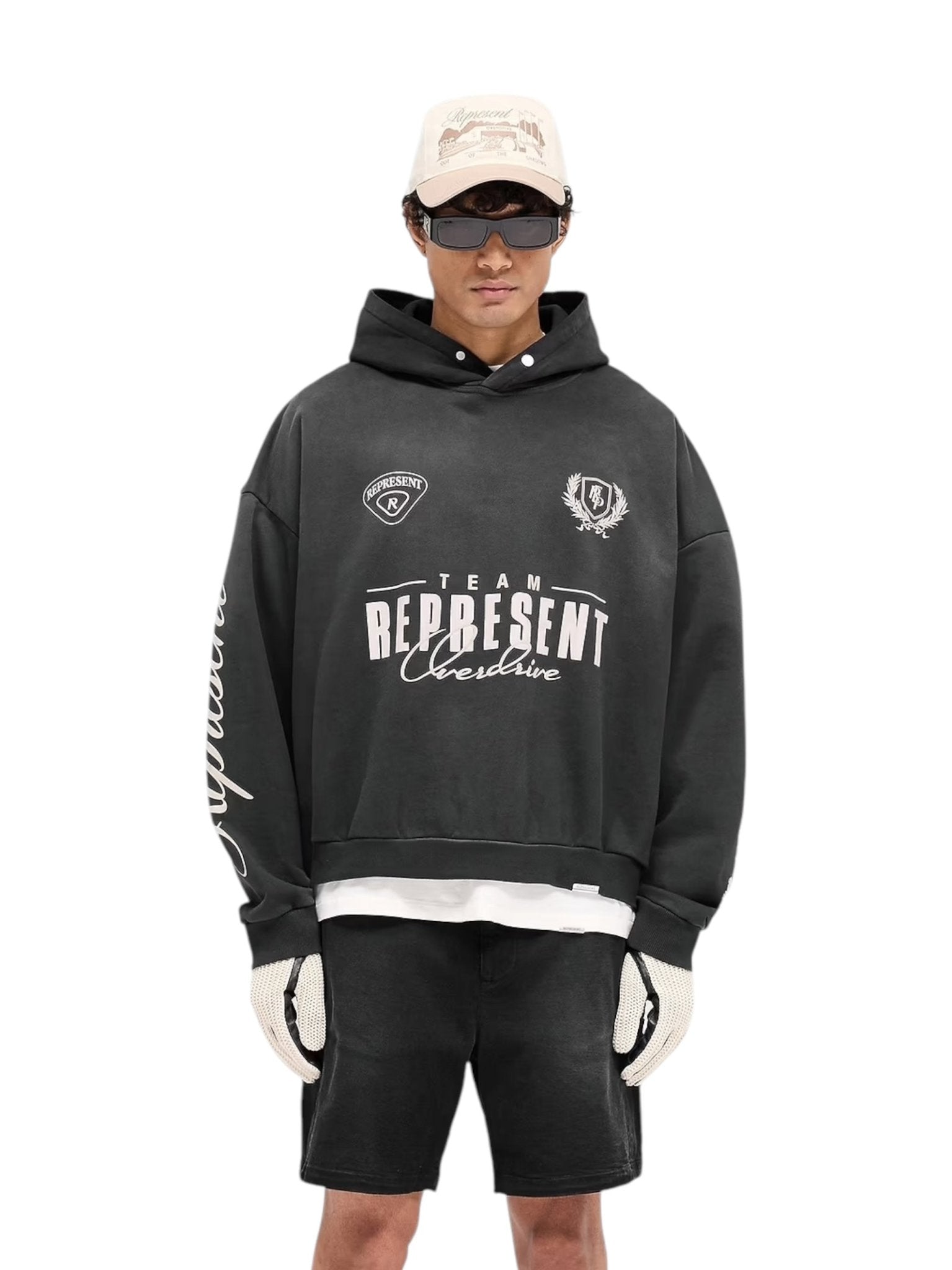 A person confidently stands against a crisp white background, wearing the Represent MLM41144 World Championship Hoodie in Black by REPRESENT, featuring an oversized boxy fit for a vintage look. They're paired with black shorts, a beige cap, sunglasses, and white gloves.